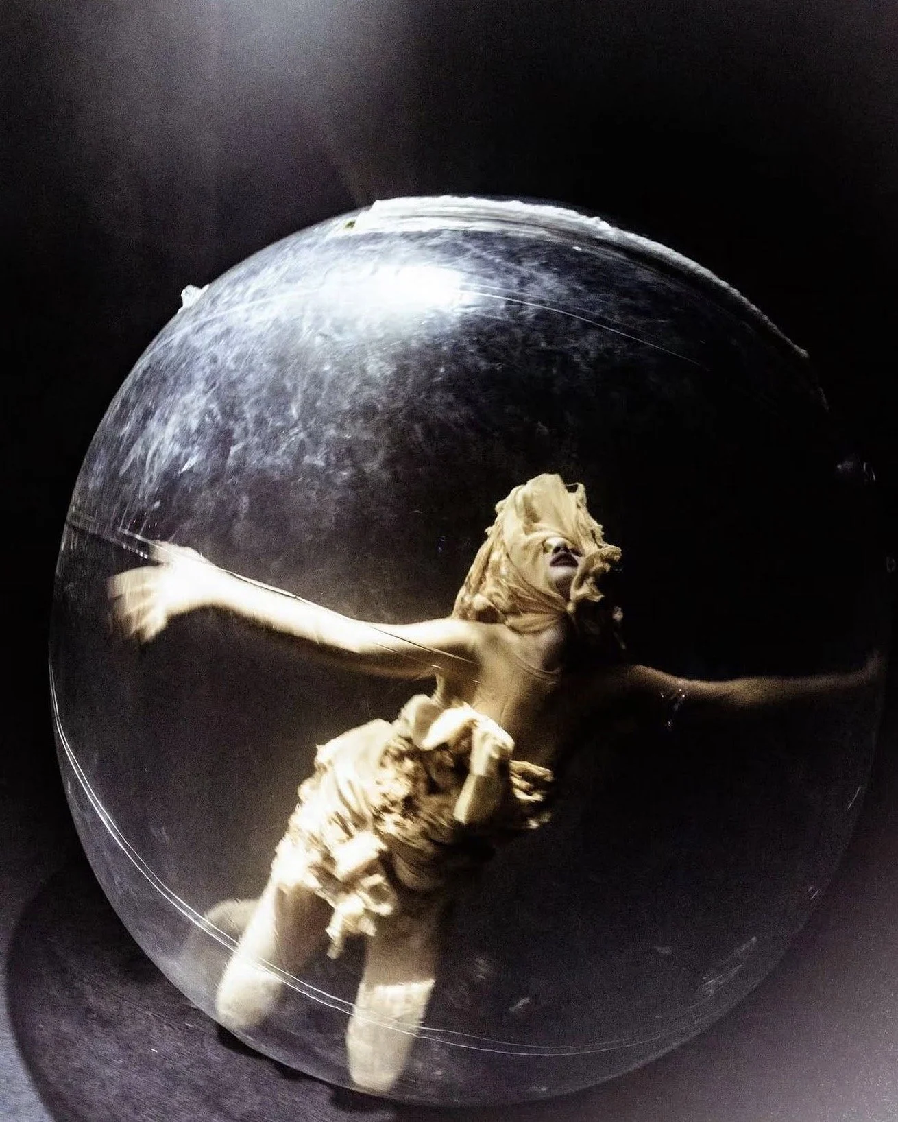 A sculpture of a woman inside a glass sphere, with arms outstretched and hair flowing, set against a dark background.