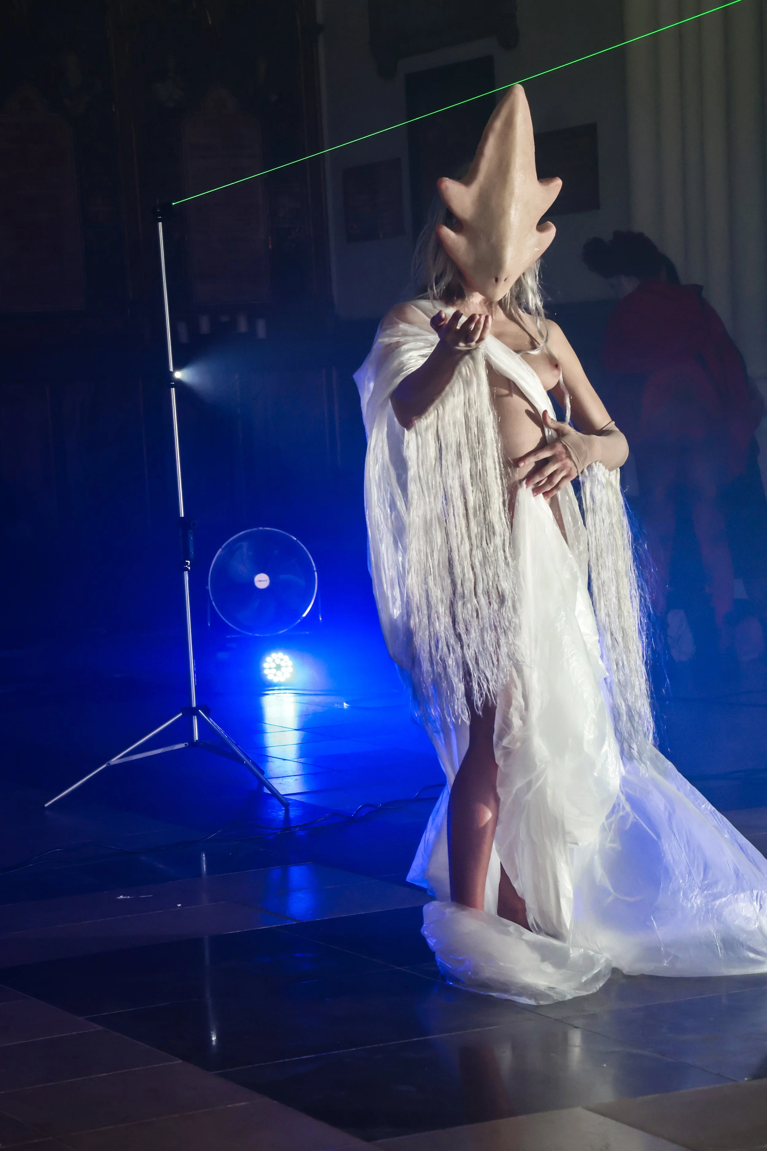 Person wearing a creature mask, draped in a flowing white garment, standing on a stage with blue lighting and a laser beam.