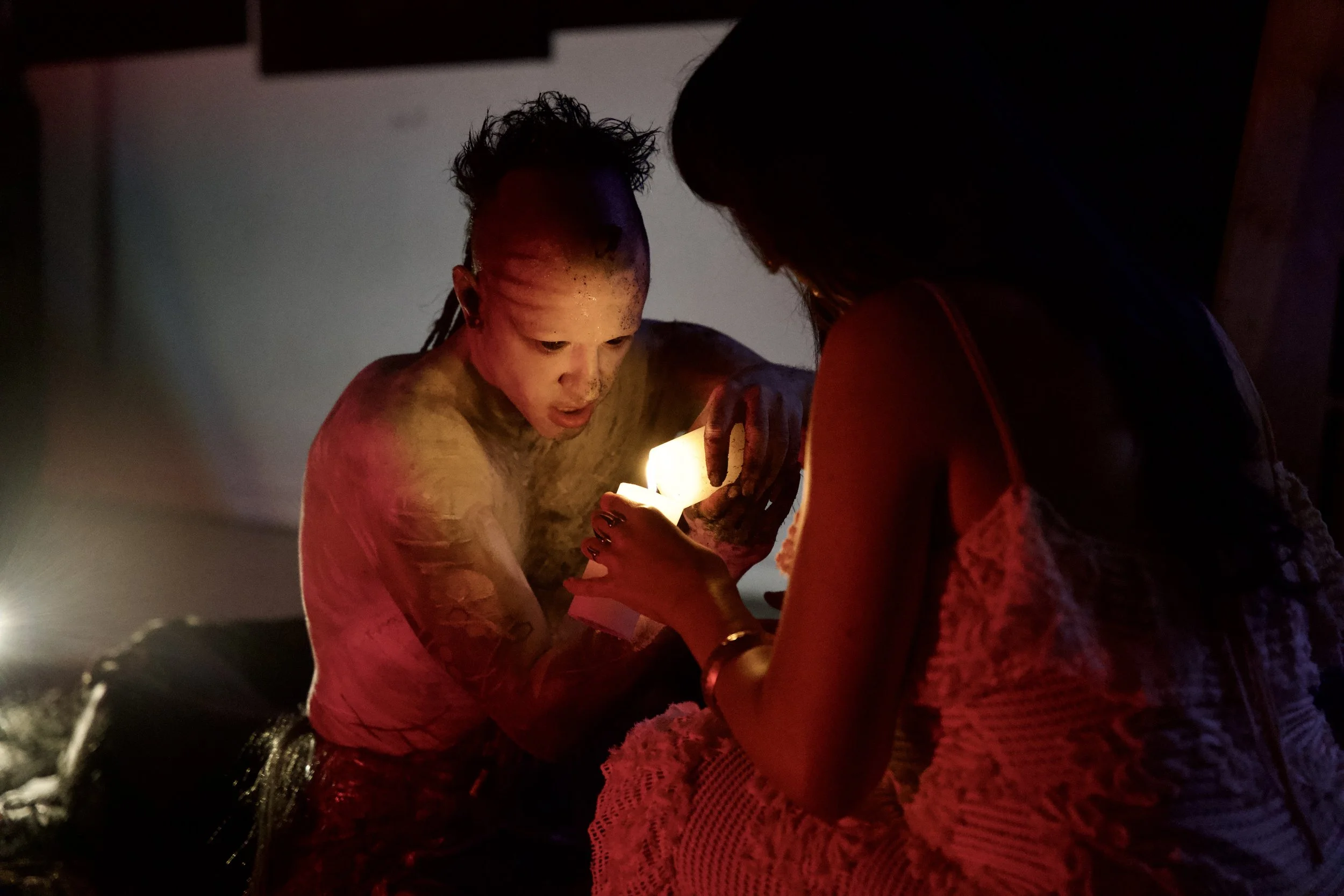Two women in a dark room illuminated by a candle, one with facial tattoos and colorful makeup, the other in a sleeveless lace top, engaging closely with the candle.