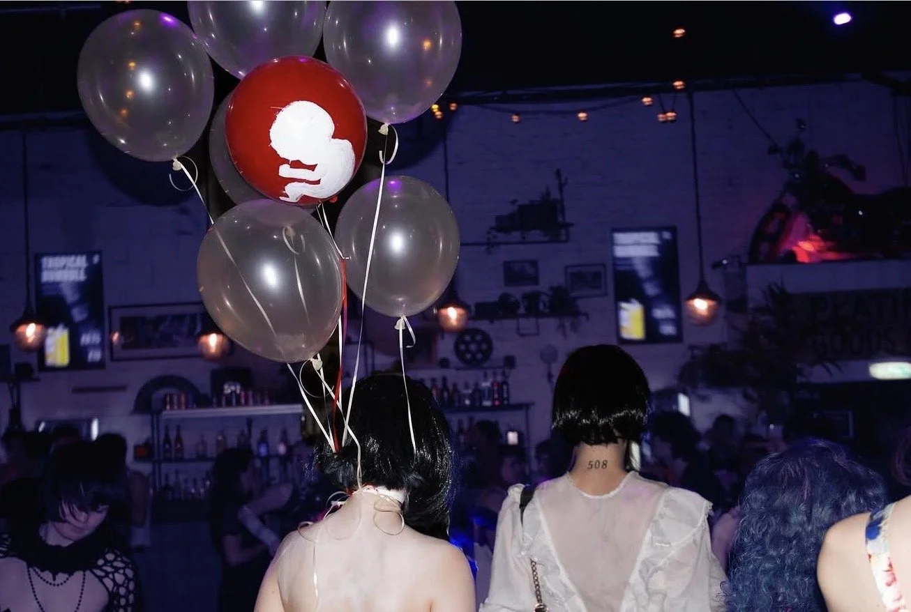 Group of people at a dimly lit party or nightclub, features balloons including a red one with white silhouette of a head in profile, in a room decorated with framed pictures and string lights, bar shelves and a TV in background.