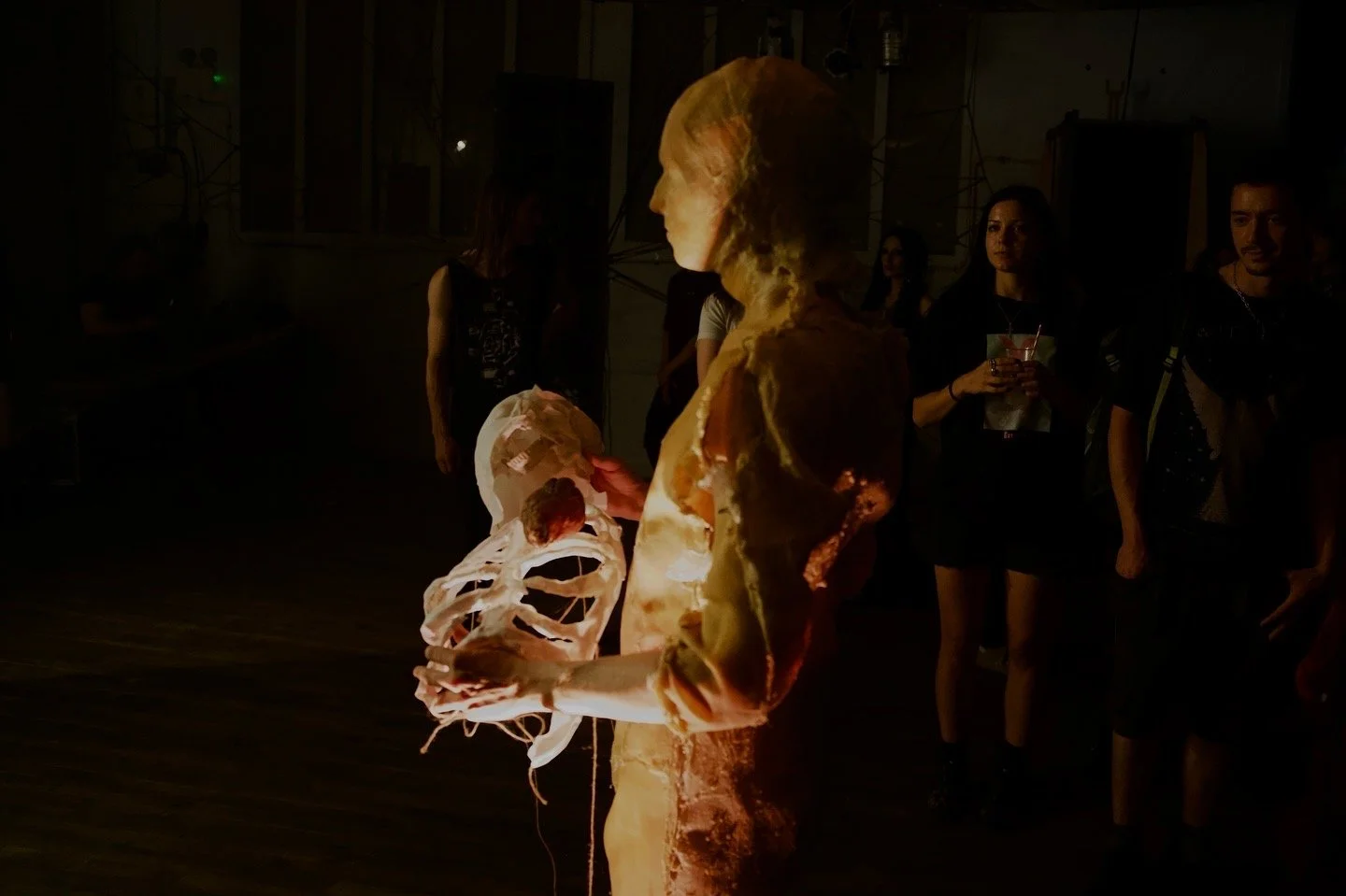 A person dressed as a human skeleton is standing in a dark room with several onlookers in the background. The skeleton costume is illuminated, showcasing a detailed ribcage and skull.