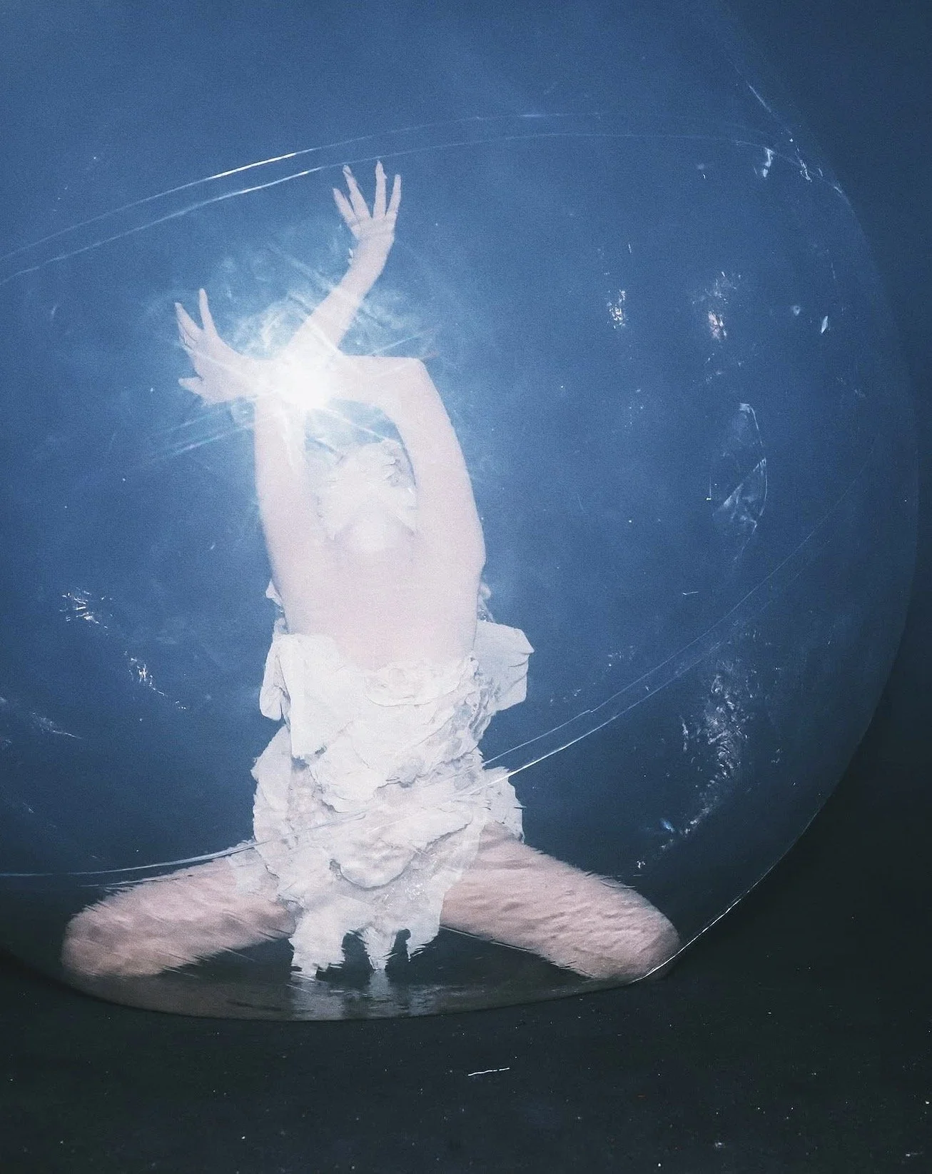 Person wearing a large transparent balloon costume, with their legs and arms visible inside, kneeling with arms raised and hands pressed against the balloon surface.