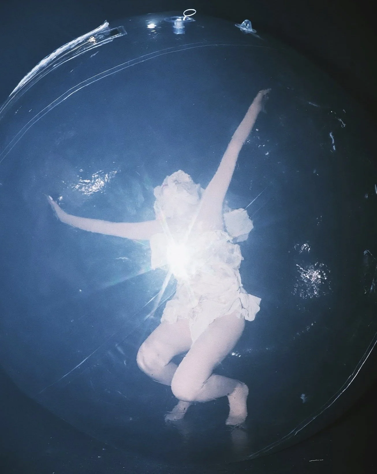 A child in a white dress and tights is underwater in a swimming pool, with arms outstretched and head tilted back, capturing a playful moment.
