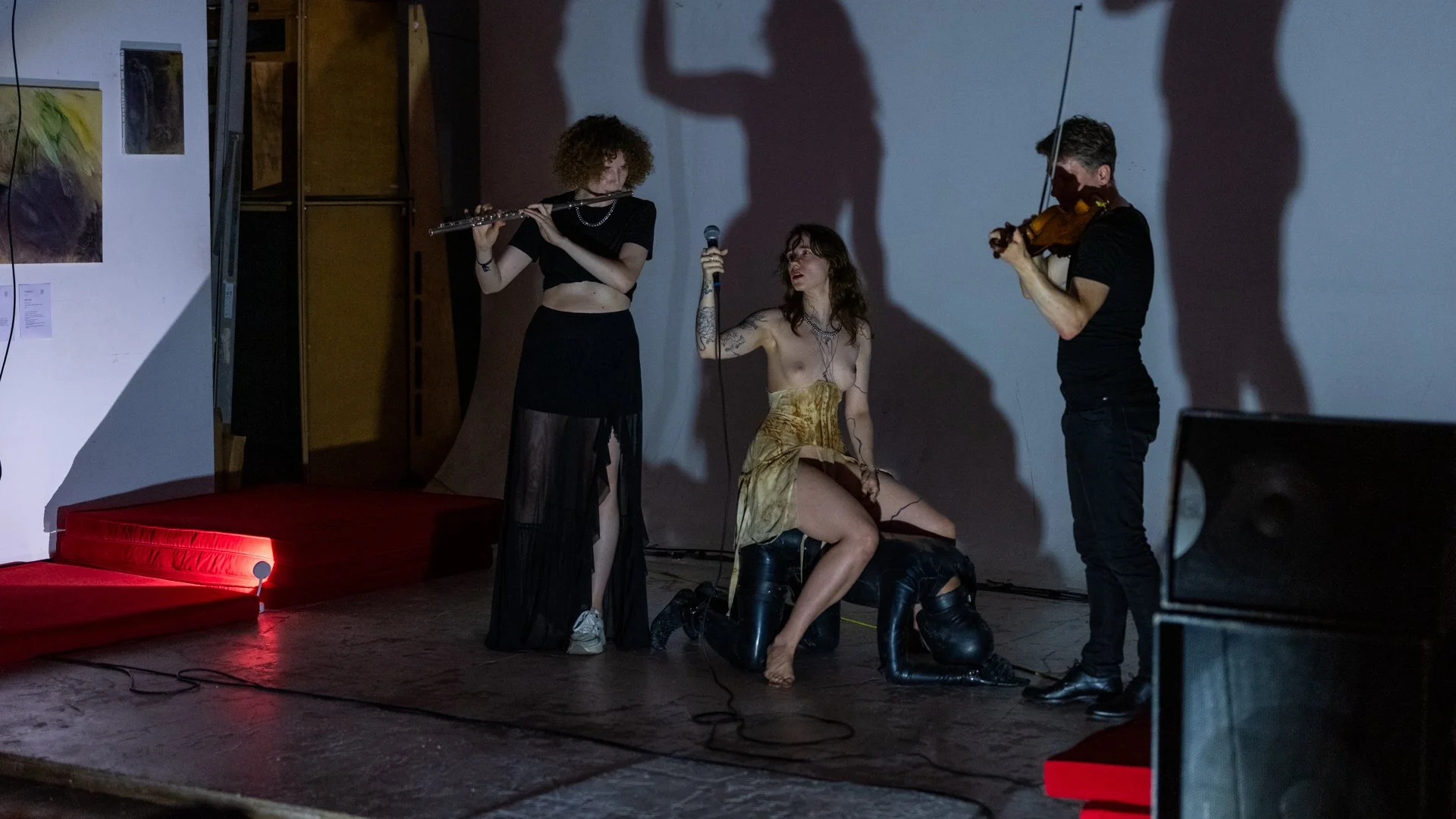 Three performers on stage with shadows cast on the wall behind them. One woman is kneeling and holding a microphone, wearing a yellow skirt, black boots, and jewelry. A woman with curly red hair is standing, playing a flute, wearing a black crop top 
