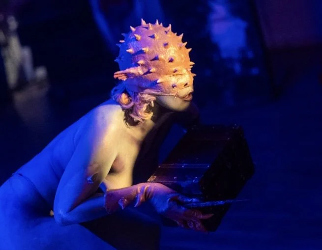 A person wearing a spiked, orange, dome-shaped mask with a face of a fish, holding a black box.