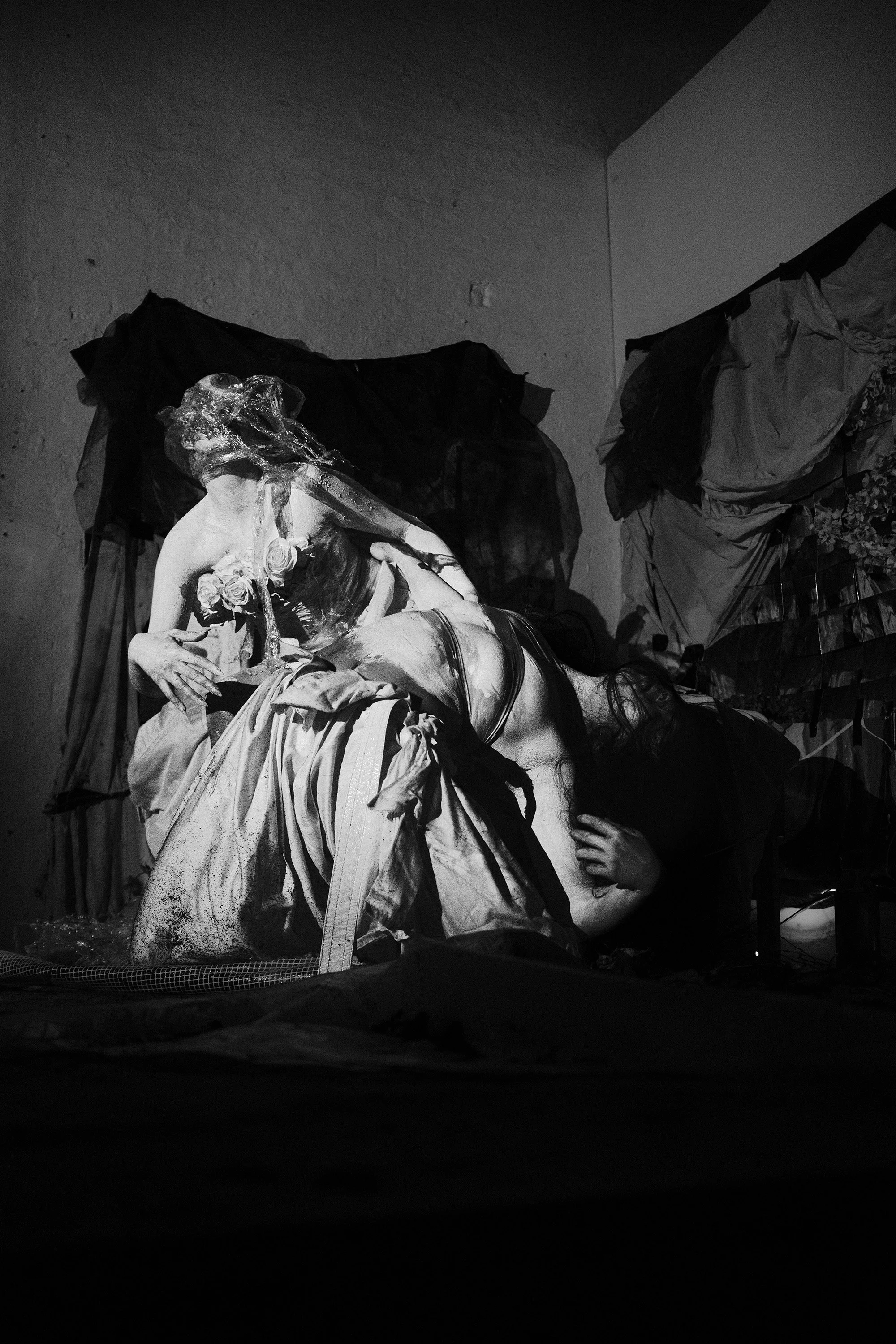 Black and white photograph of a sculpture of a woman holding a bouquet of roses, with a veil over her face, and another person leaning on her leg in a dimly lit indoor setting.