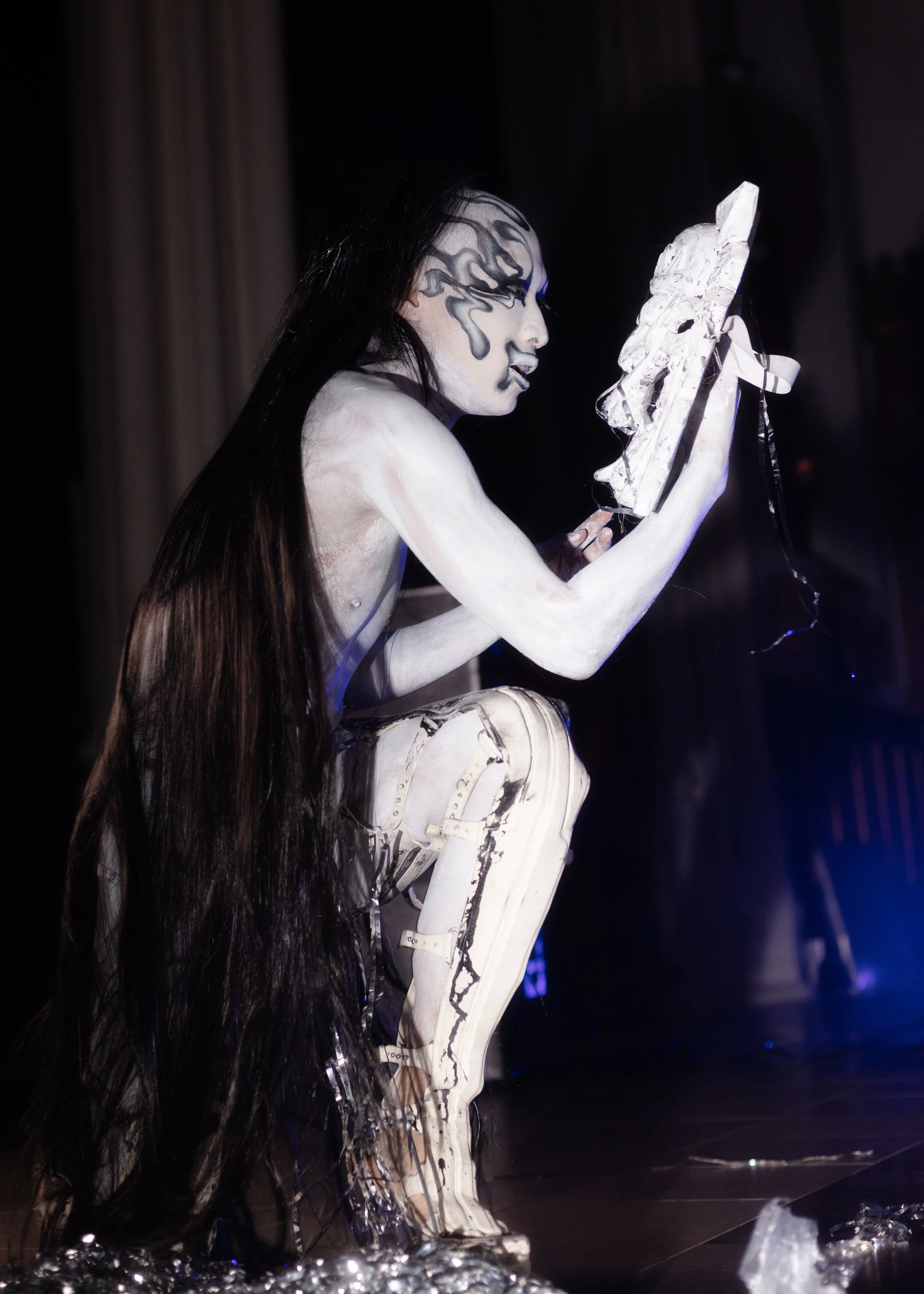 Person with long black hair and white body paint, crouching and holding a large, white, sculptural object, on a dark stage with curtains in the background.