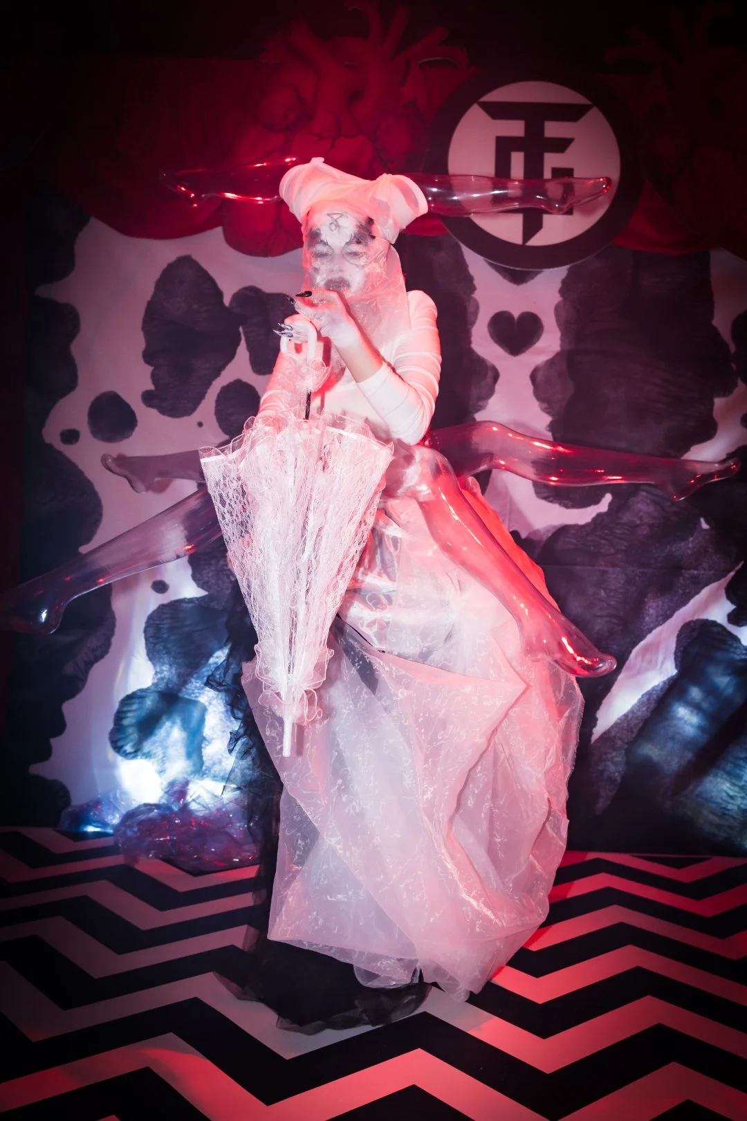 A person dressed in avant-garde fashion with a white and pink layered dress and a headpiece, holding an umbrella, in a stylized, theatrical setting with red and black lighting and a patterned floor.