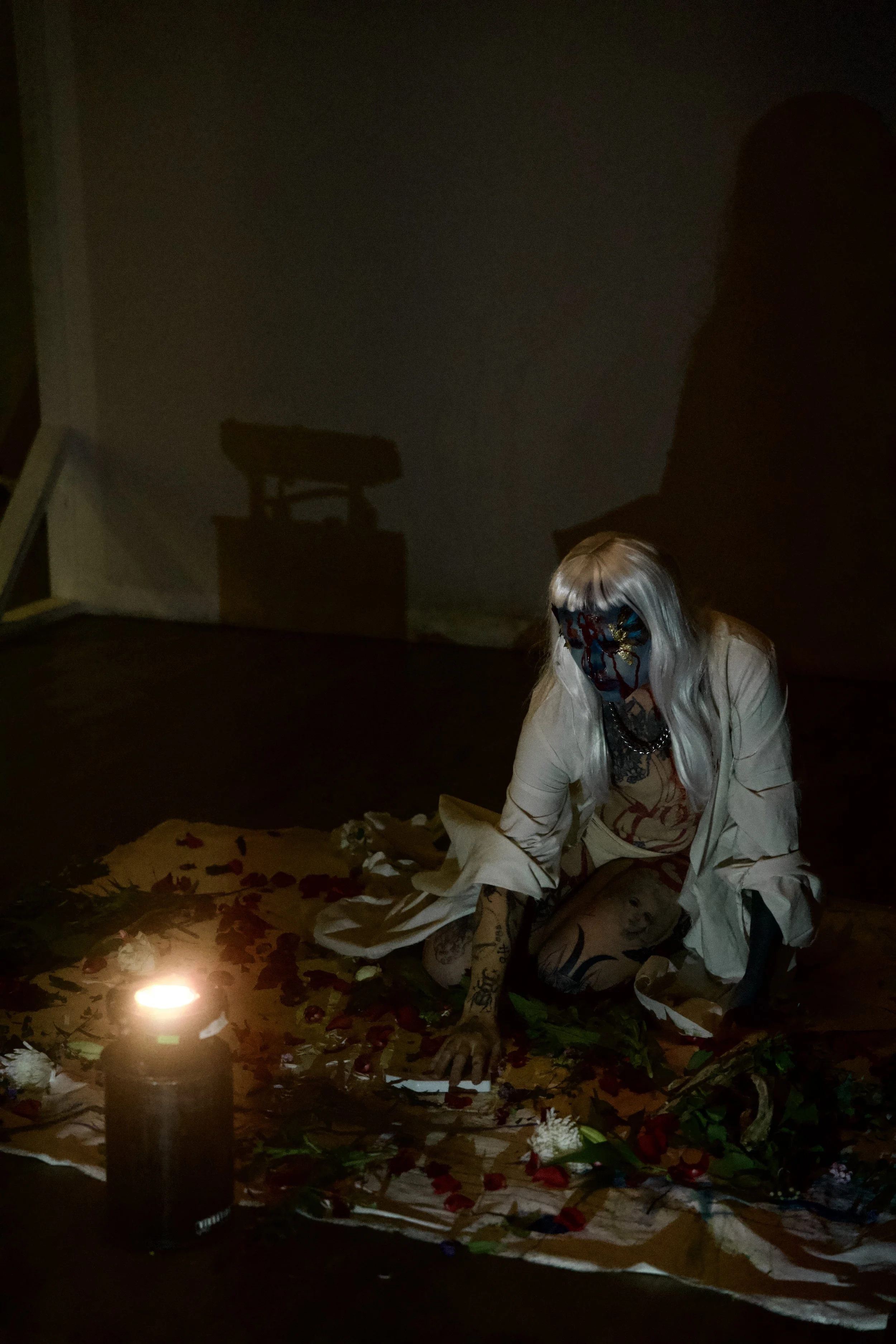 Person with long silver hair and colorful makeup, dressed in white, kneeling on the floor surrounded by flower petals and greenery, illuminated by a single candle, in a dark room.