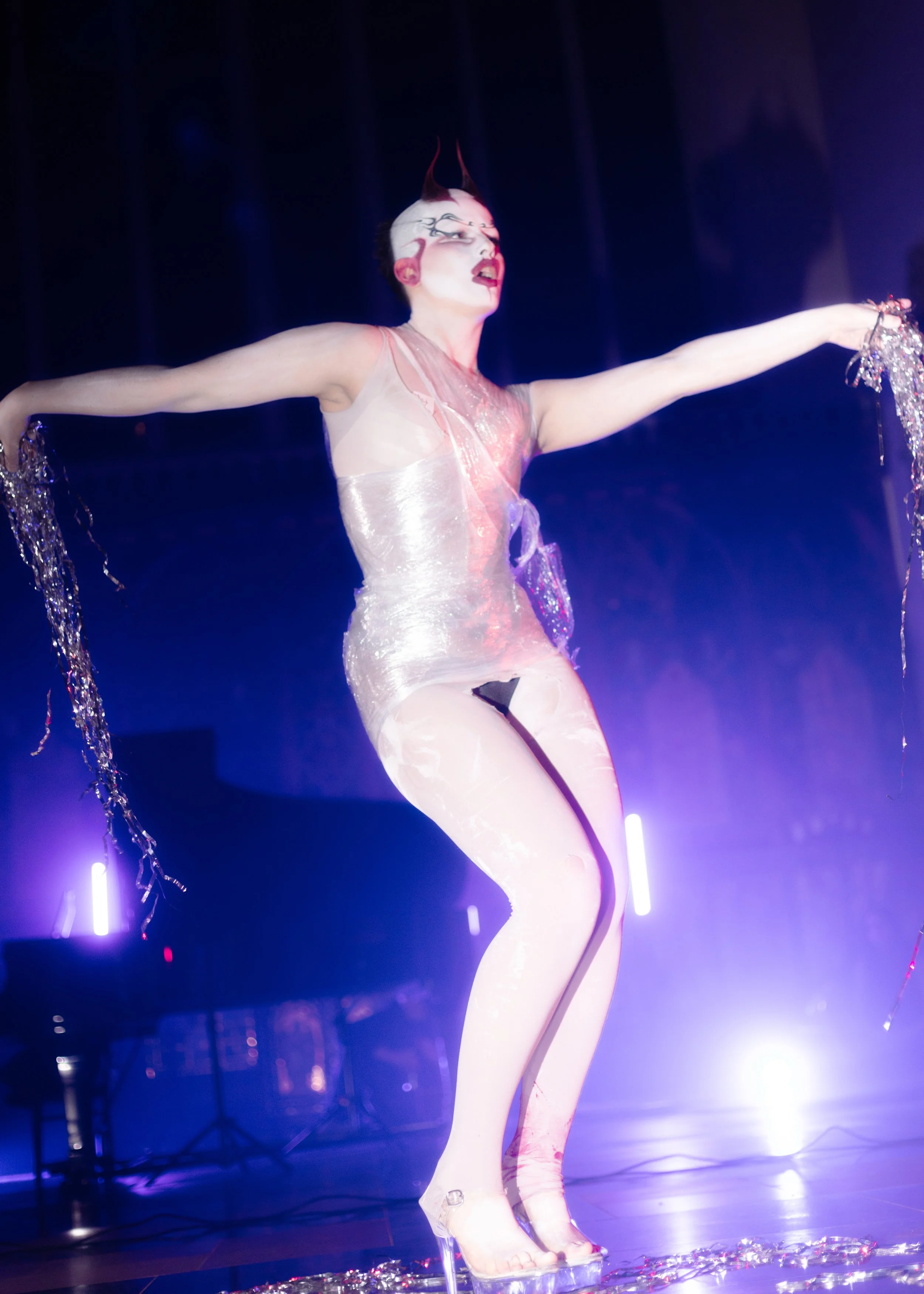 Performer on stage with white face paint, red lipstick, and horns, wearing a shiny, sheer outfit with high heels, holding metallic streamers in both hands, lit by stage lights.