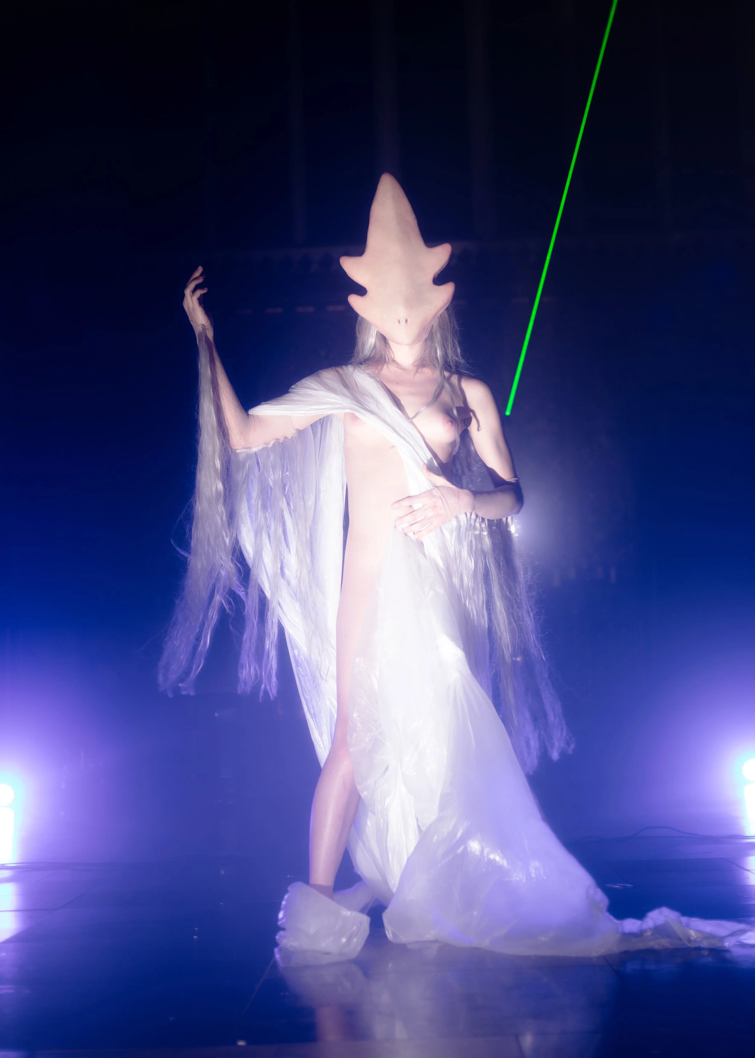 A person wearing an elaborate costume with a shark-shaped headpiece, flowing white fabric, and long blonde hair, standing on stage under bright lights with a dark background.