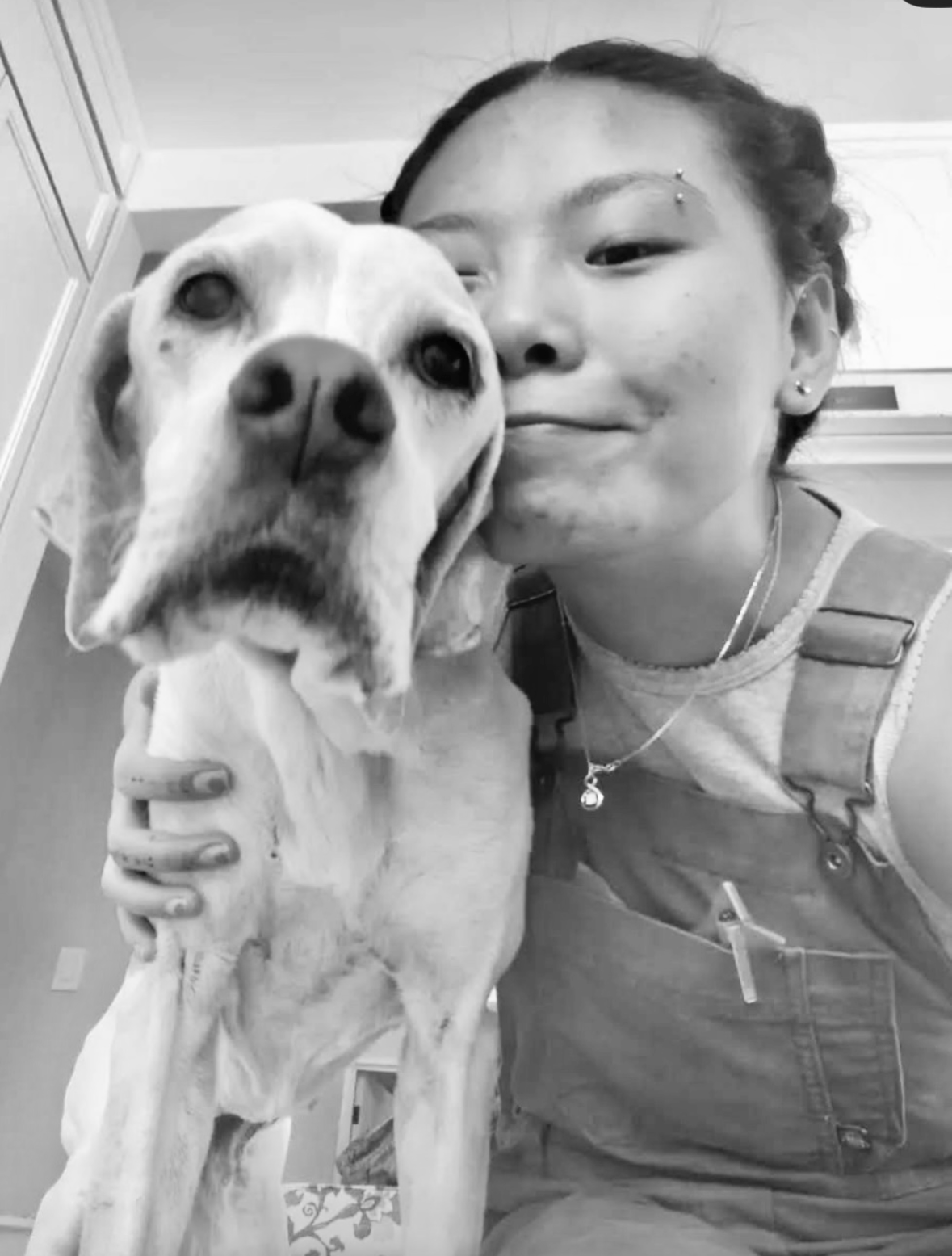 Black and white photo of a young woman with black hair and multiple facial piercings, smiling while holding a large dog with light fur, floppy ears, and an open mouth, in an indoor setting.