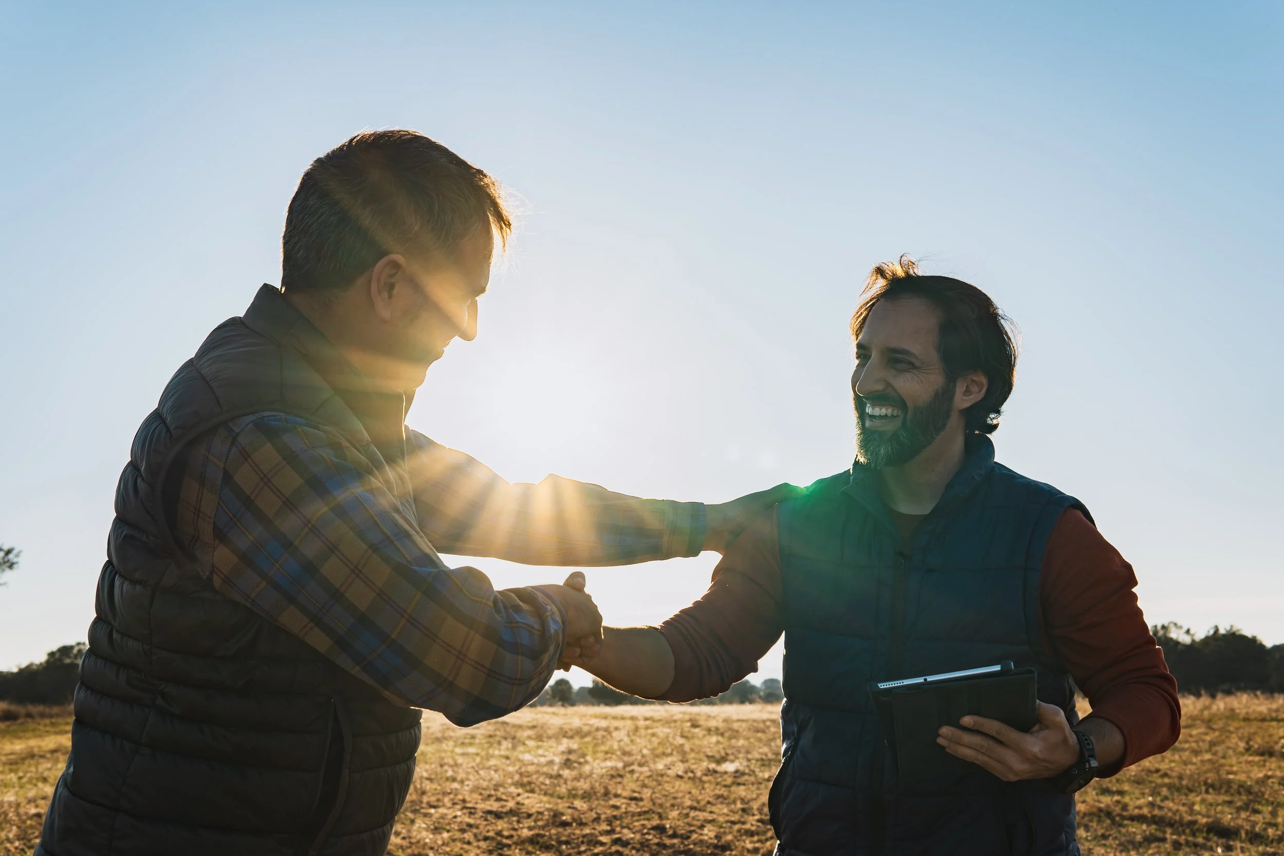 Two men shake hands outdoors in a field under a clear blue sky, with one holding a tablet, as the sun sets or rises casting a warm glow.