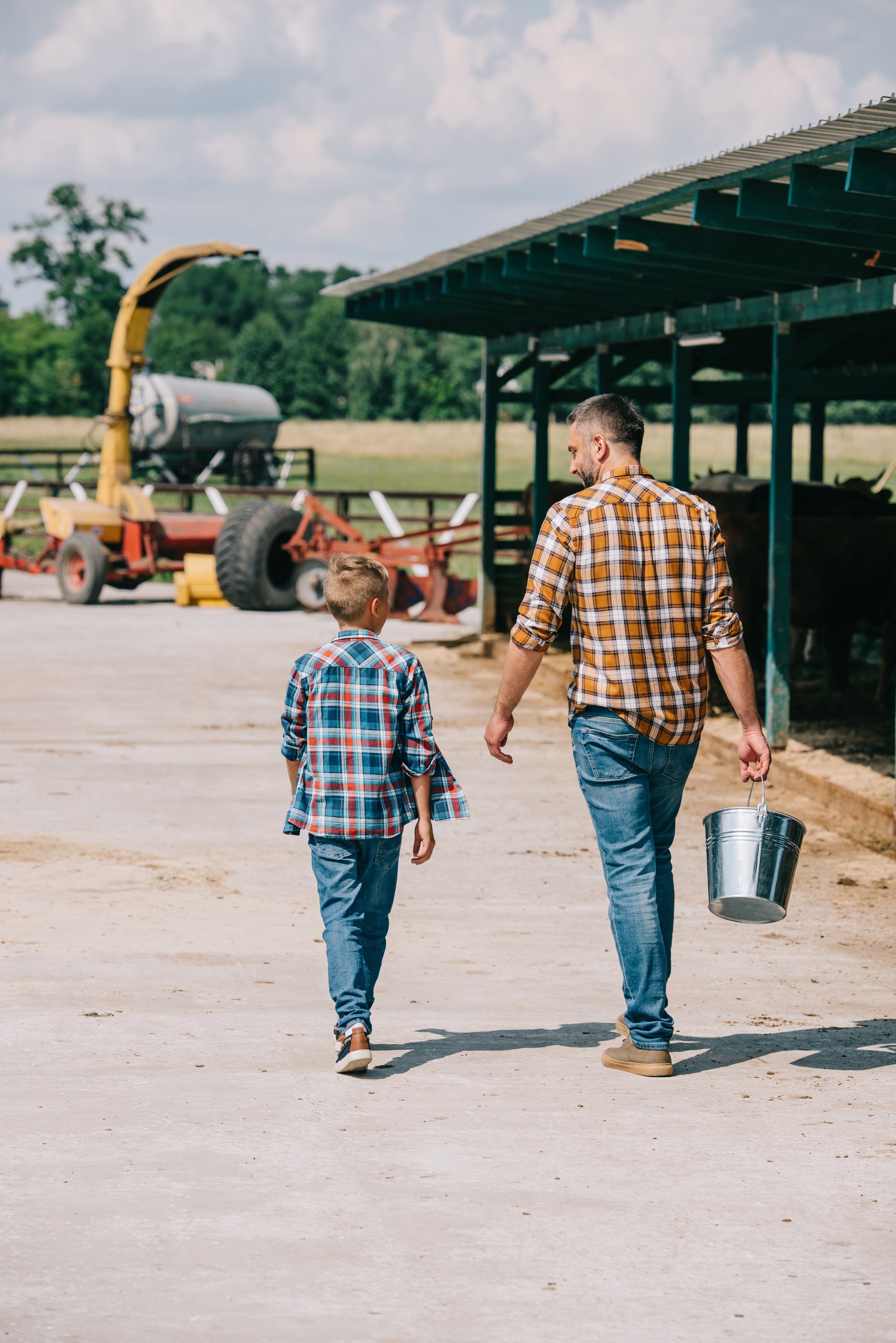 A man and a young boy walking on a farm, with a barn and farming equipment in the background, both wearing plaid shirts.