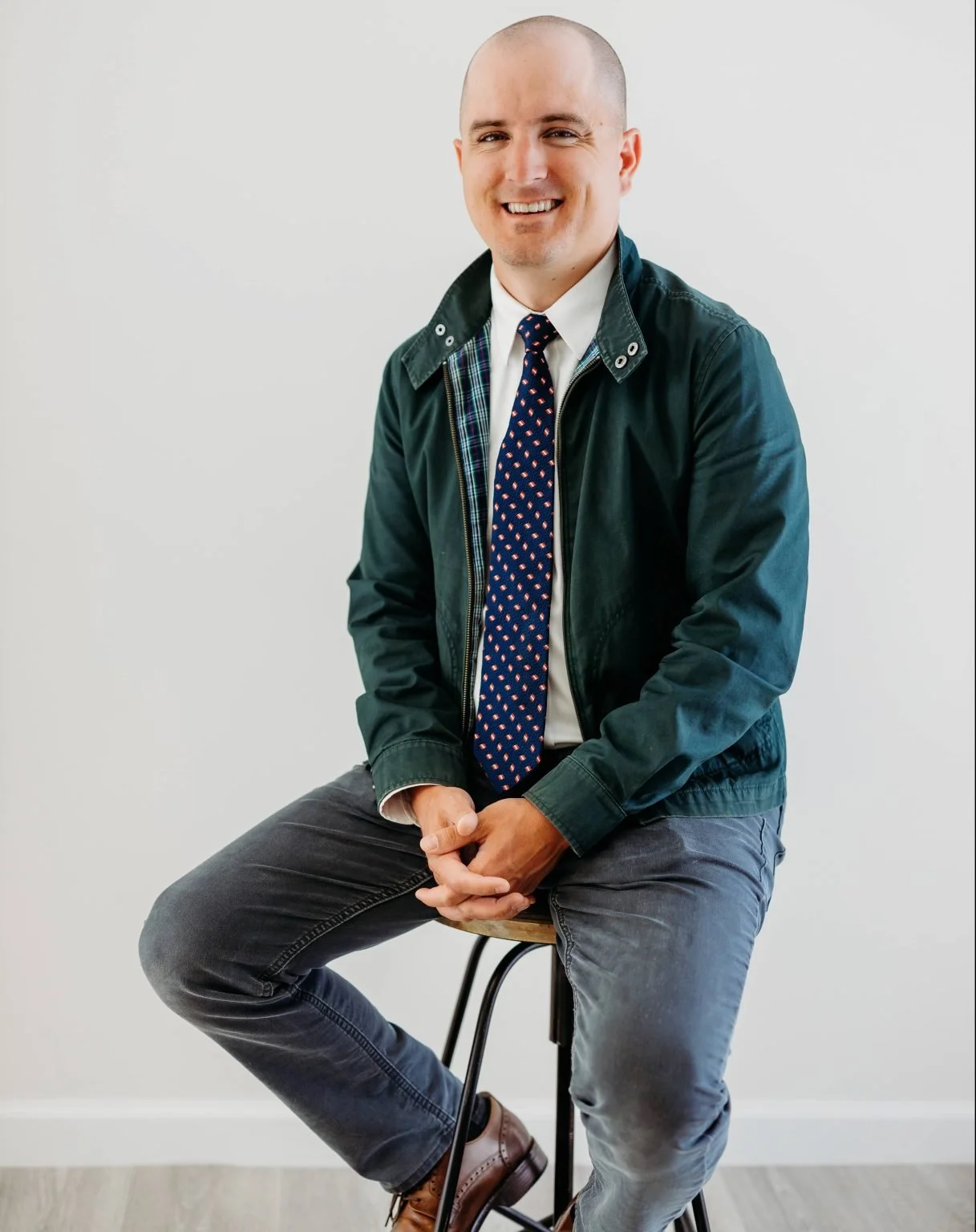 A smiling man with a shaved head sitting on a stool against a white wall, wearing a dark green jacket, a white shirt, a patterned tie, gray jeans, and brown shoes.