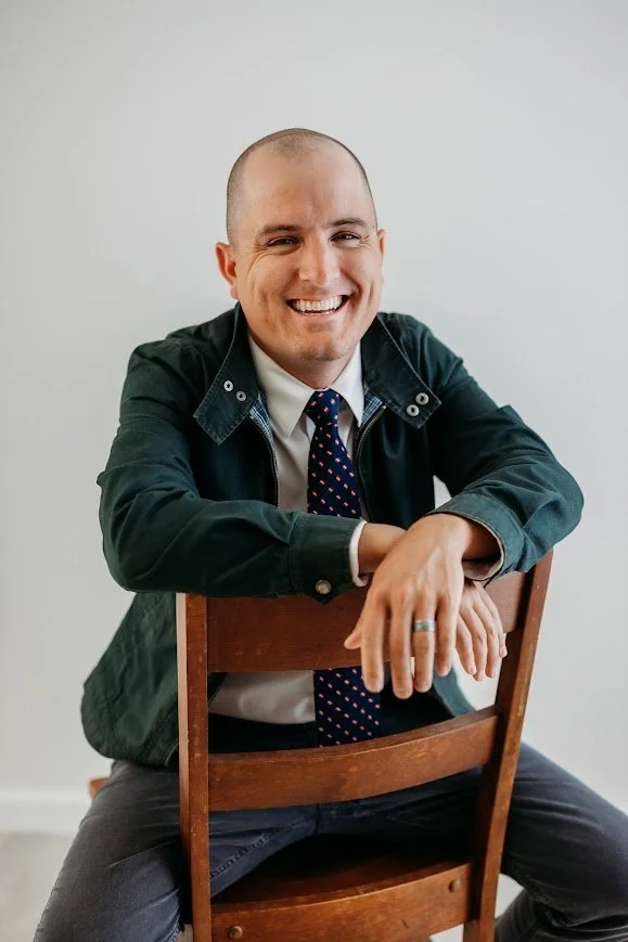 A man with a bald head and a big smile sitting on a wooden chair, leaning forward with his arms resting on the back of the chair, wearing a dark jacket, a white shirt, and a navy blue tie with red dots, against a plain light-colored background.