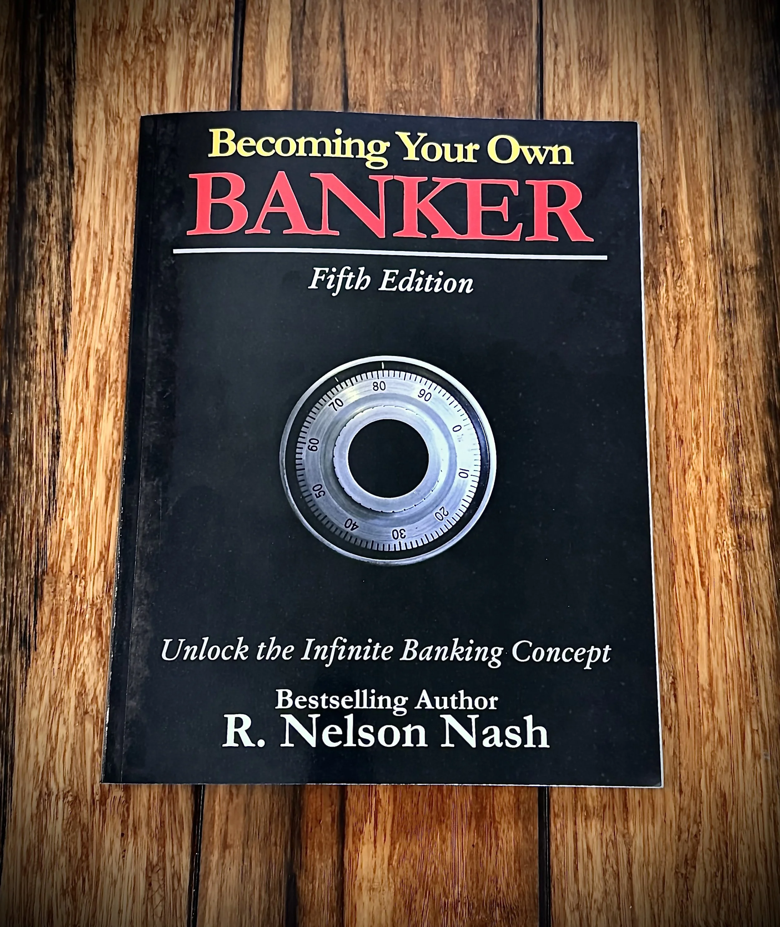 Book cover titled 'Becoming Your Own Banker, Fifth Edition' by R. Nelson Nash, placed on a wooden surface.