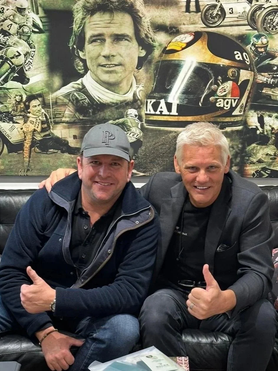 Two men sitting on a black leather couch smiling and giving thumbs up, with a racing-themed collage mural behind them featuring a race car helmet, portraits of race car drivers, racing motorcycles, and a race car.