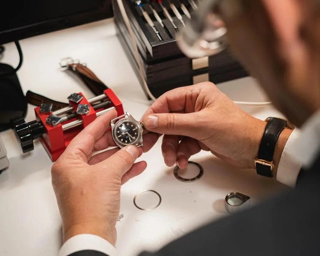 A person working on a watch, inspecting it closely in a workshop with watchmaking tools and parts on the table.