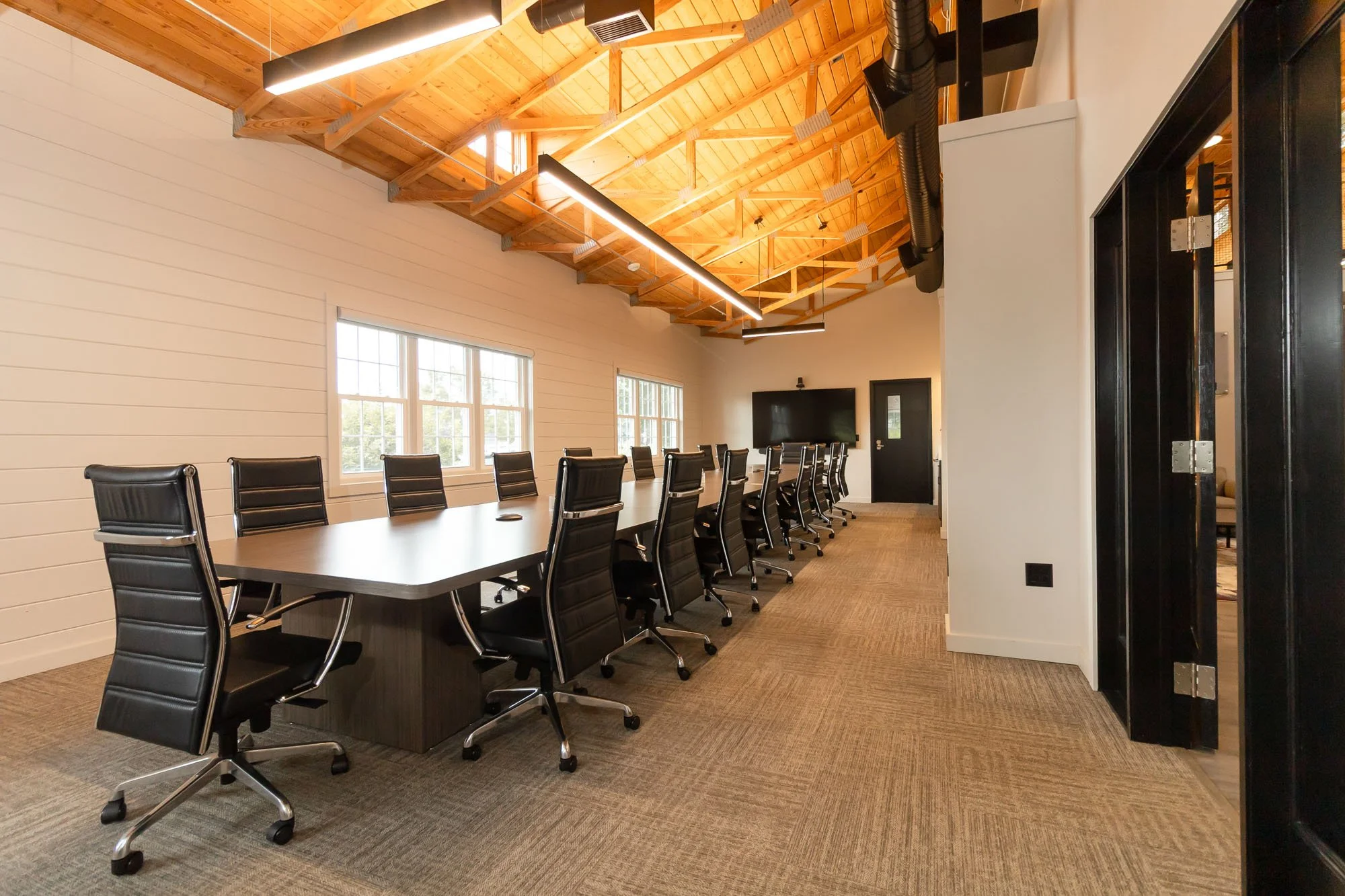Modern conference room with a long table surrounded by black leather chairs, large windows letting in natural light, a wood vaulted ceiling with exposed beams, and a large wall-mounted TV.