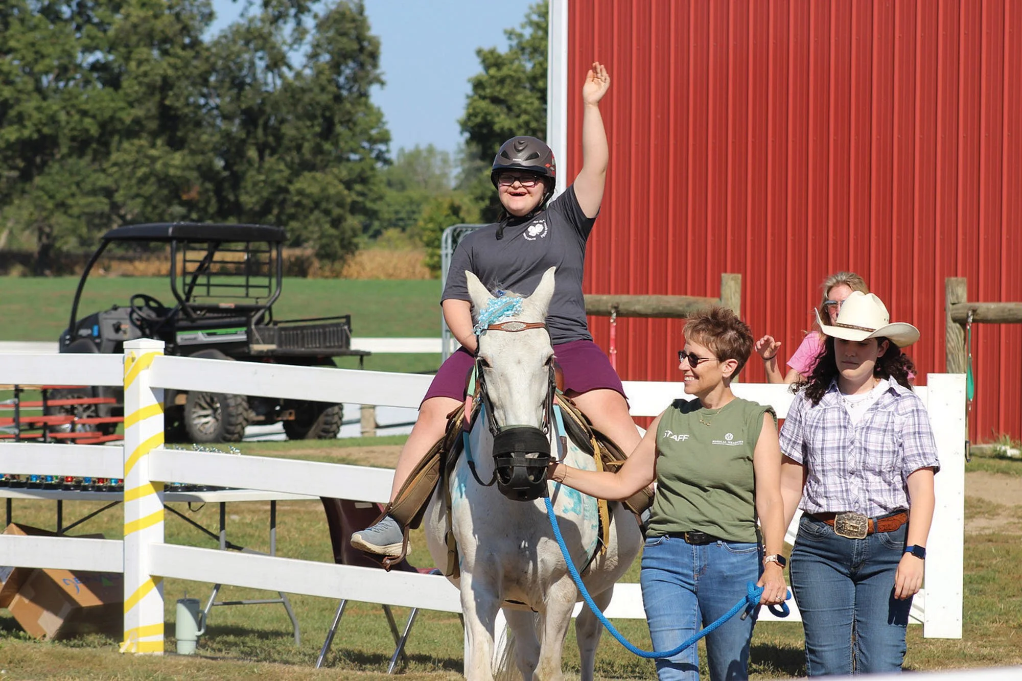 A young girl wearing a helmet is riding a white horse over a small obstacle at an outdoor event, with adults and a red barn in the background.