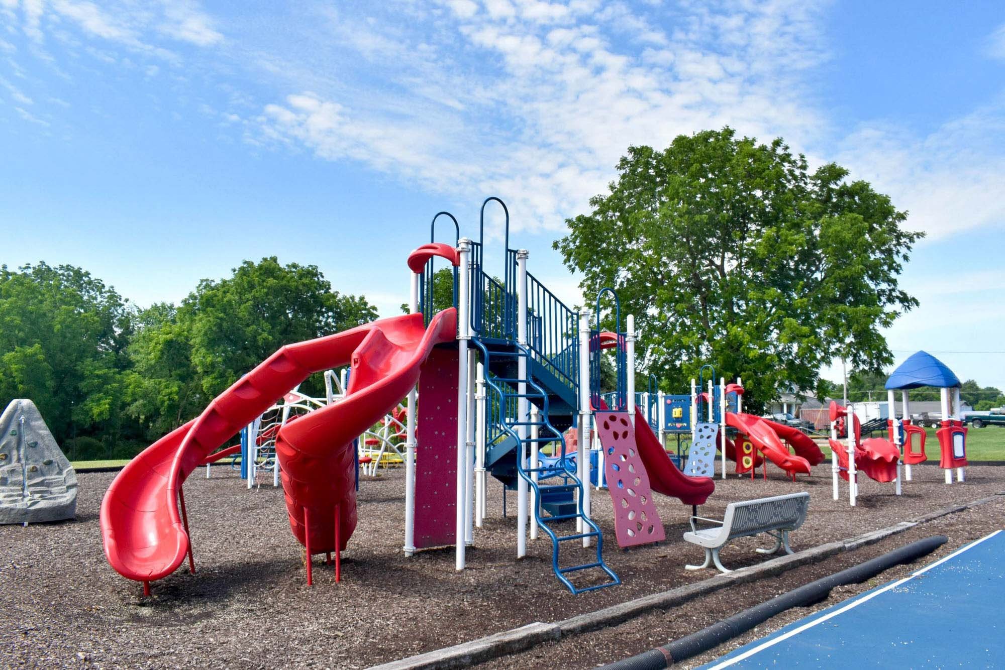 Colorful playground with slides, climbing structures, and benches, set against green trees and a bright blue sky with scattered clouds.