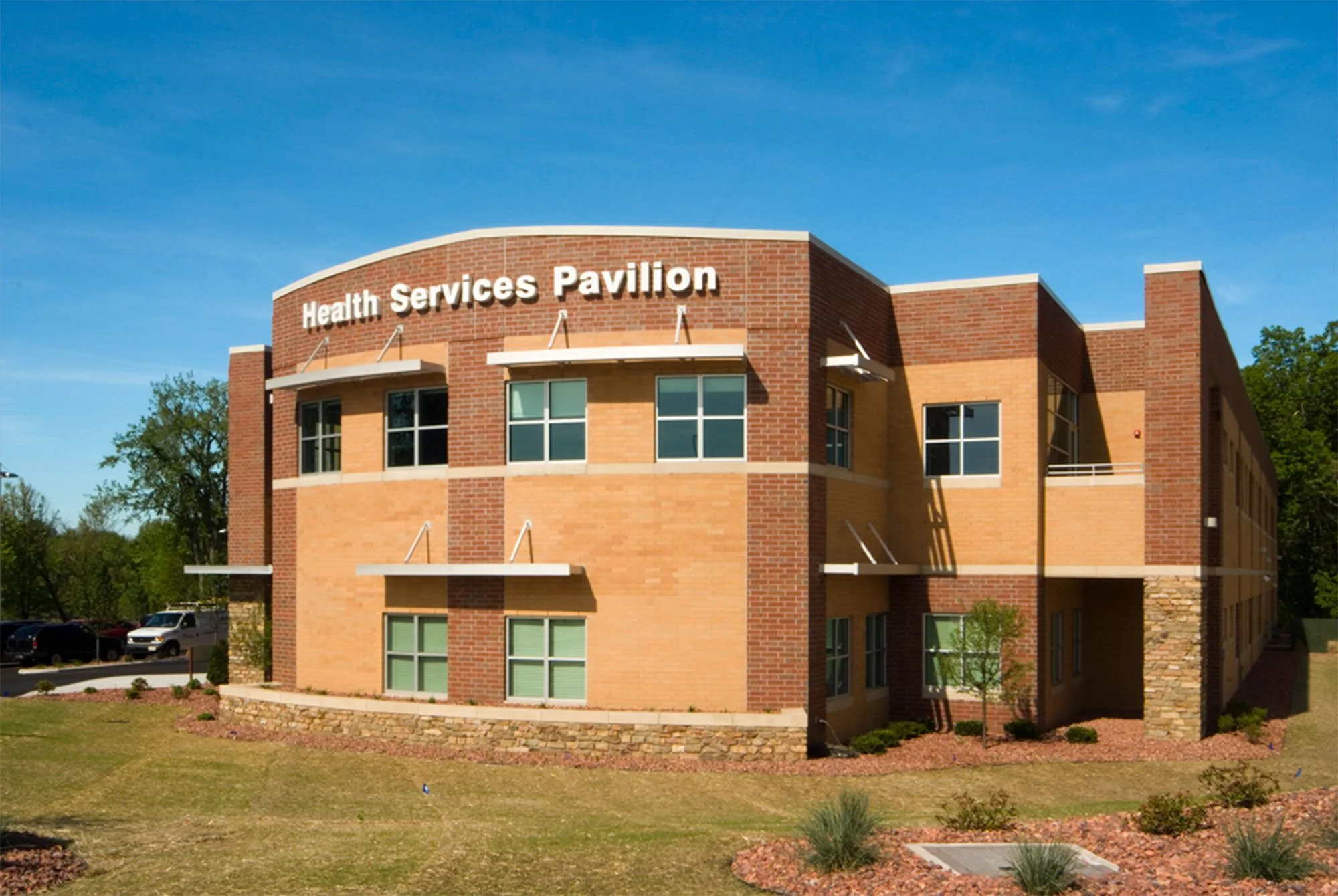 The image depicts a modern brick building with a sign that reads 'Health Services Pavilion' under a clear blue sky with some trees in the background.