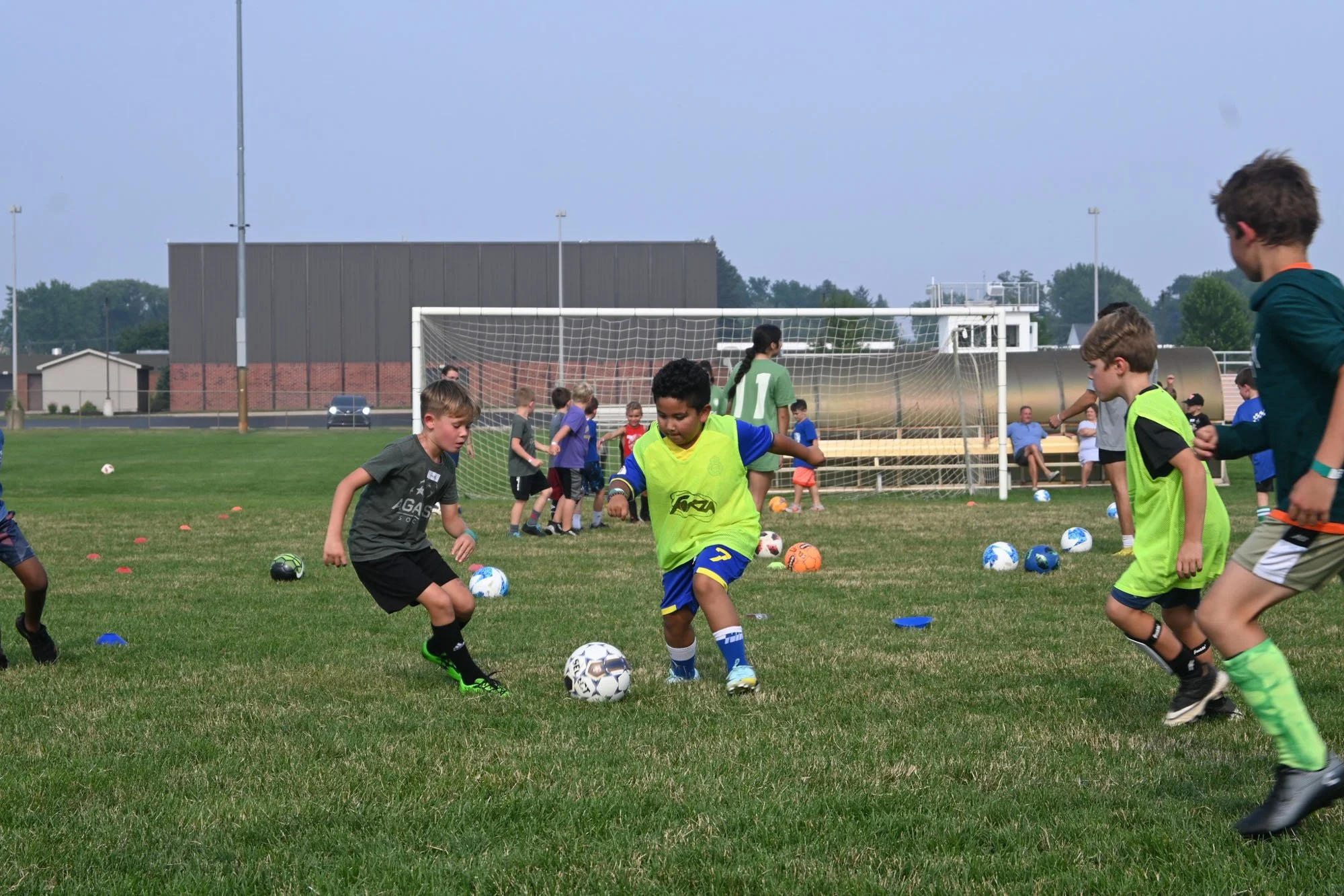 Group of children practicing soccer on a field with soccer balls, some wearing yellow training vests, a goalpost in the background, and several adults sitting on benches watching.
