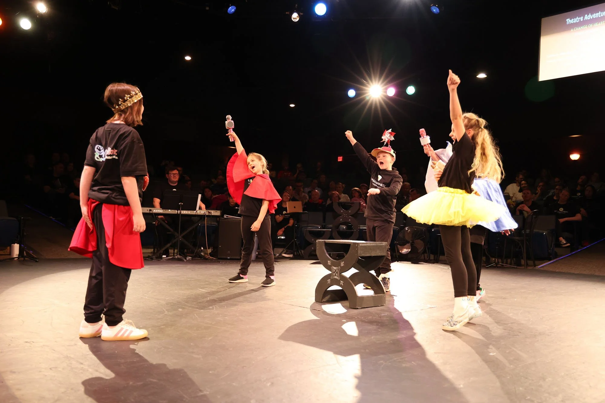 Children performing a dance routine on stage with an adult instructor, in colorful costumes and accessories, in front of an audience and stage lights.