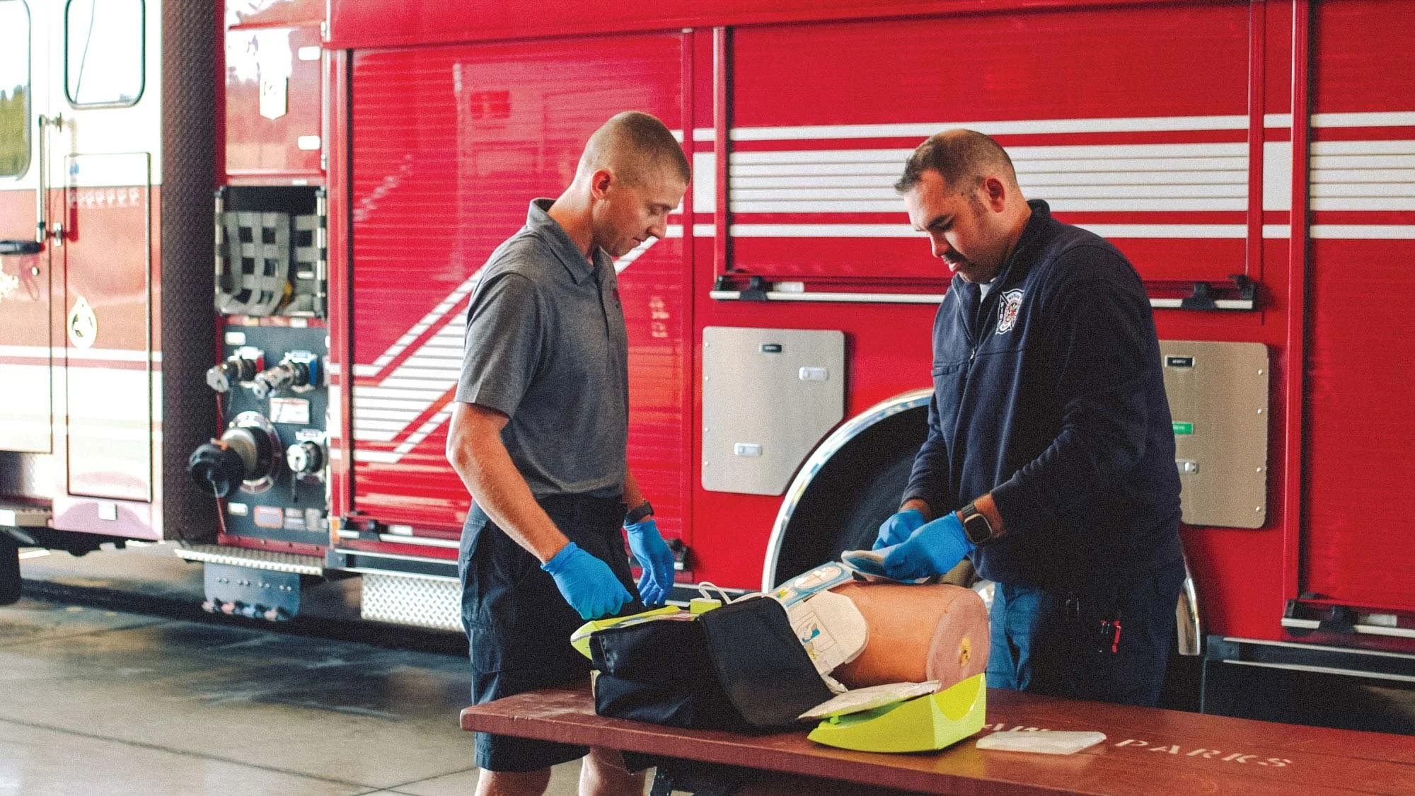 Two emergency responders, a man and a woman, wearing blue gloves and examining equipment on a table in front of a red fire truck.