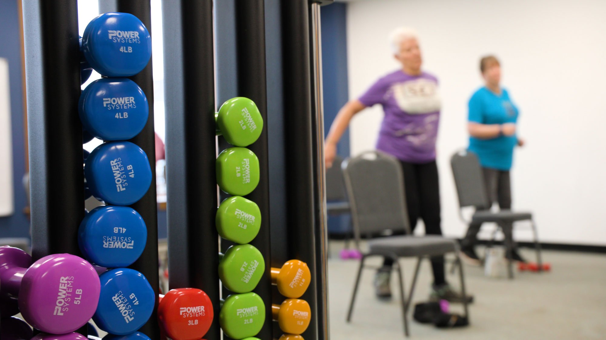 Colorful weights on a rack in a gym, with two women exercising in the background, one wearing a purple shirt and the other wearing a blue shirt.