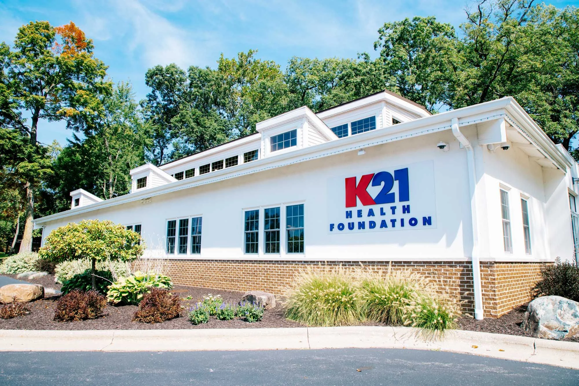 White building with K21 Health Foundation sign, surrounded by landscaped garden and trees, under a partly cloudy sky.