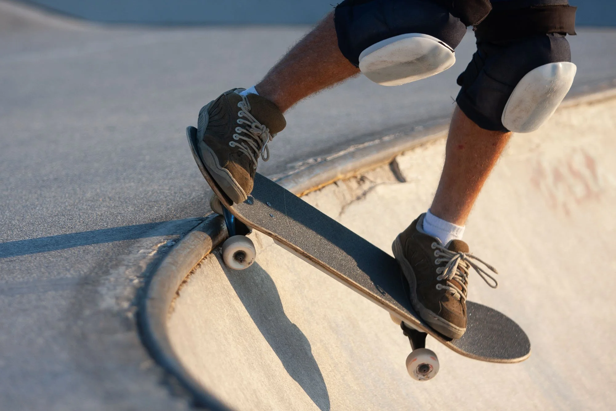 Person with brown shoes and black shorts bending down on skateboard on concrete surface, sunlight casting shadow.