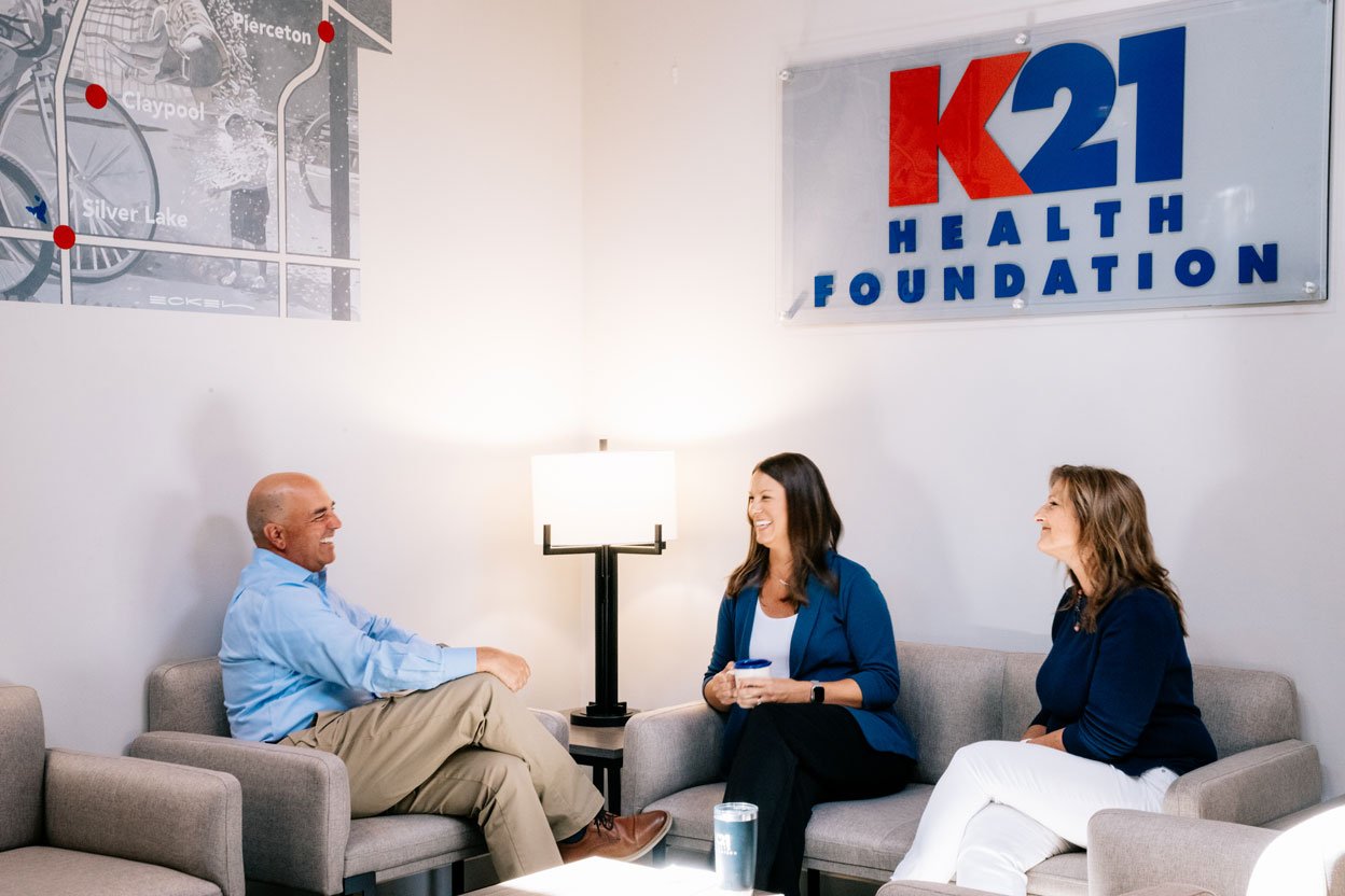 K21 Staff seated on a couch around a table talking and laughing. Large logo on the white wall behind them reads "K21 Health Foundation"