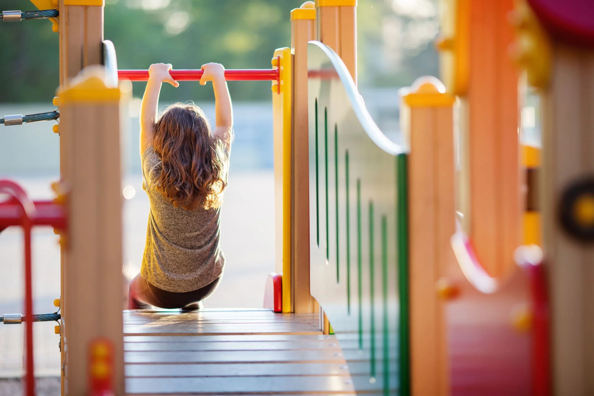 A child with long curly hair playing on a colorful playground structure, viewed from behind at sunset.