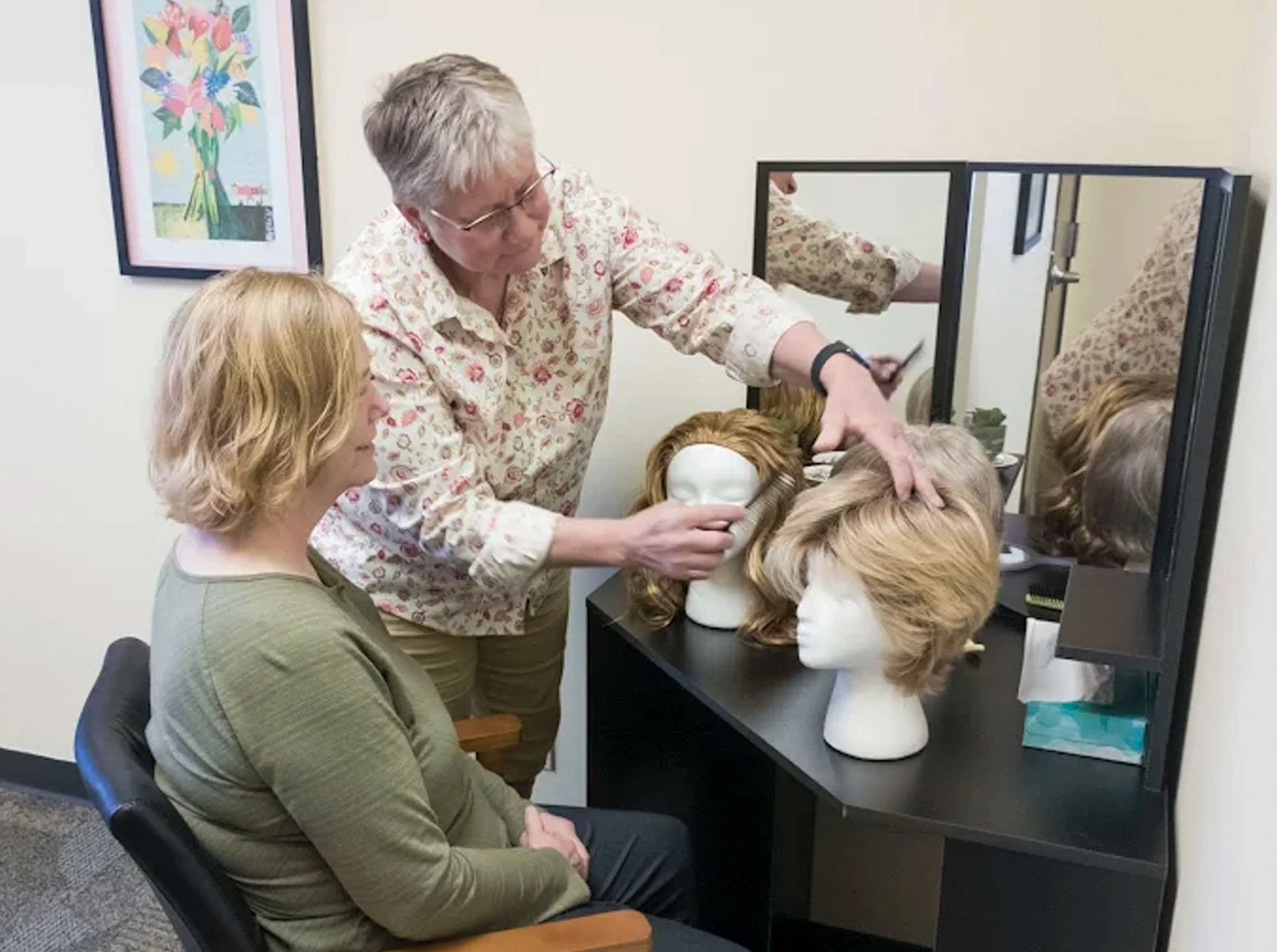 A group of women sitting and watching a hair styling demonstration with a mannequin head, a mirror, and a video camera setup.