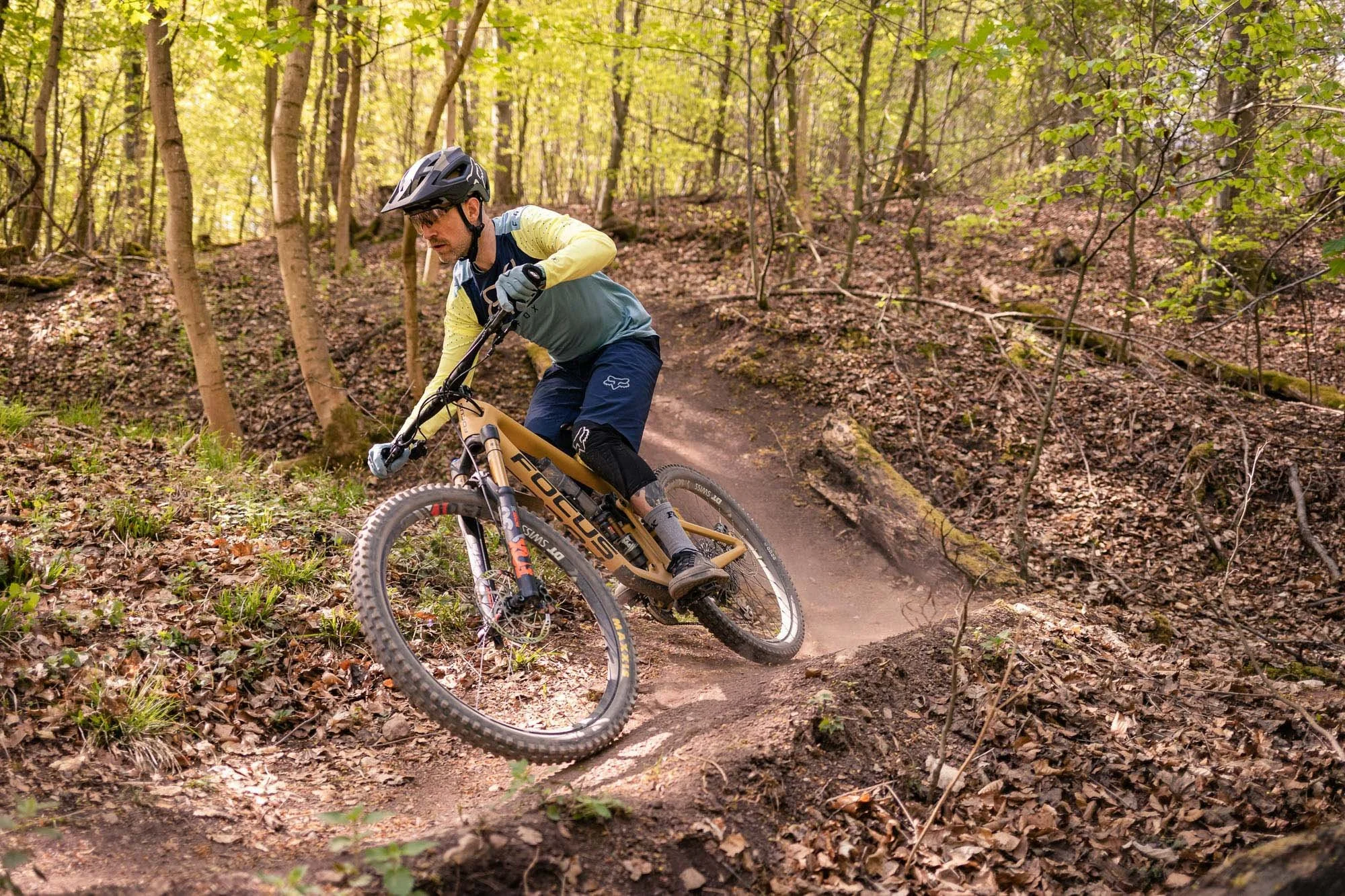 A mountain biker wearing a helmet and protective gear rides down a wooded trail with a dirt trail and green foliage.