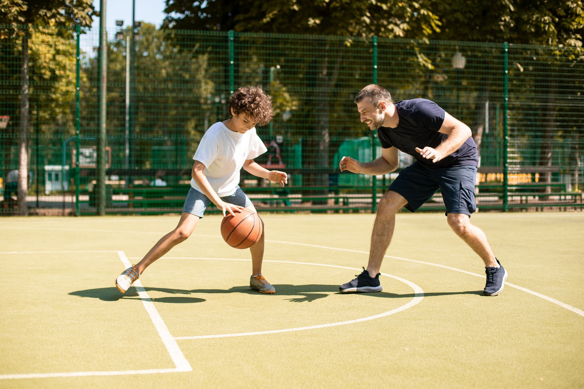A man and a young girl playing basketball on an outdoor court surrounded by a fence and trees.