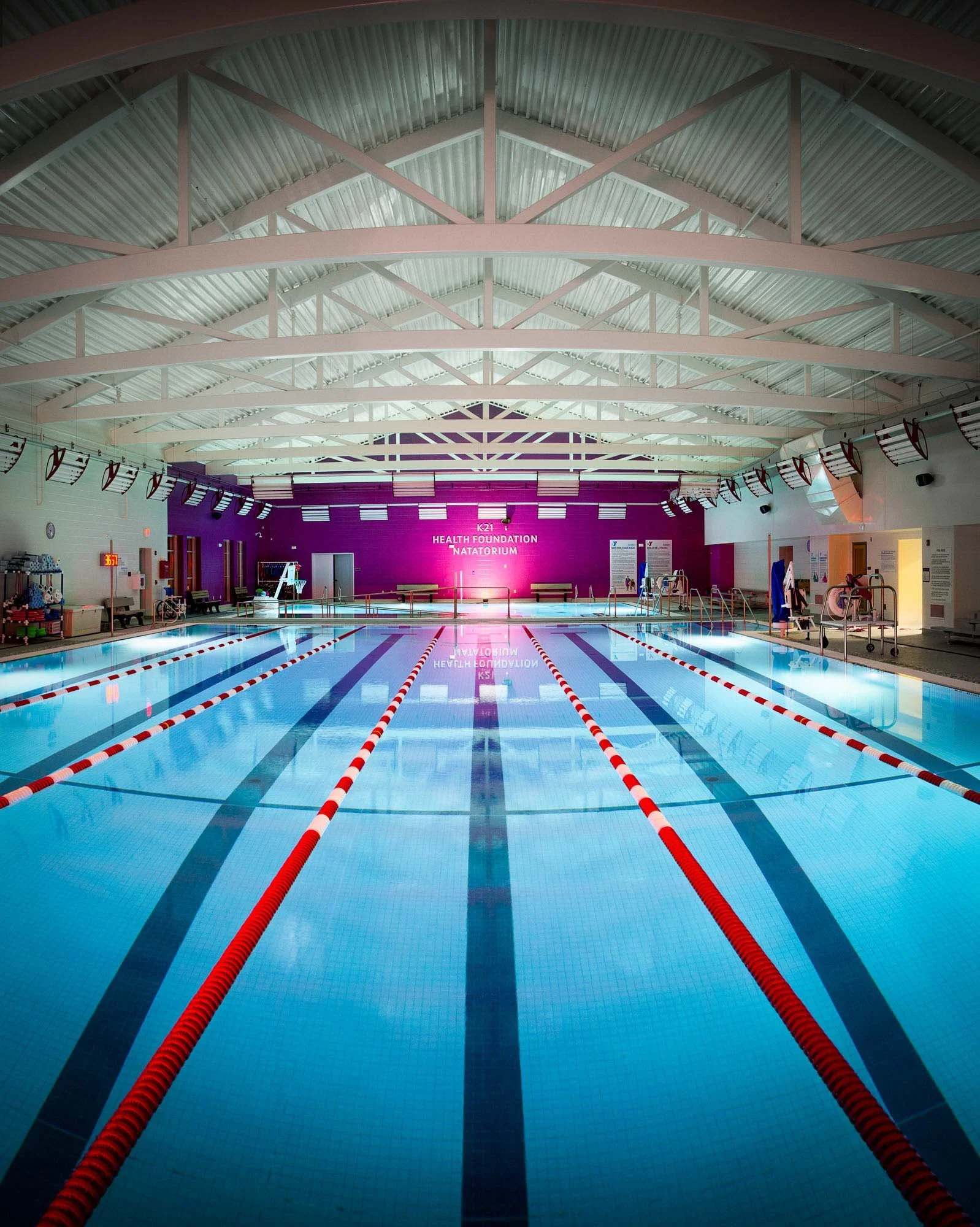 Indoor swimming pool with lane dividers, surrounded by benches, equipment, and purple walls