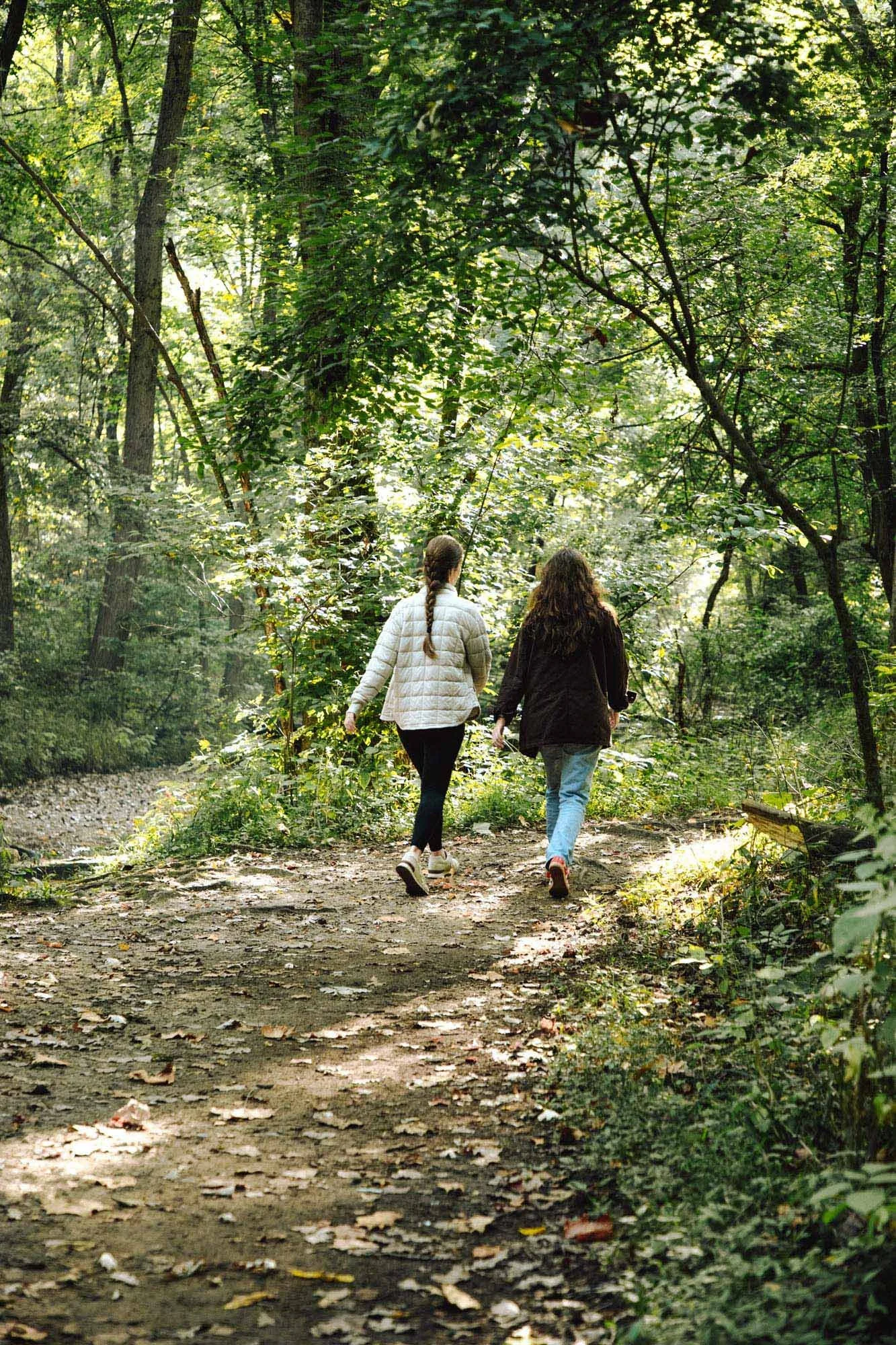 Two women walking on a dirt trail through a forest with tall trees and green foliage.