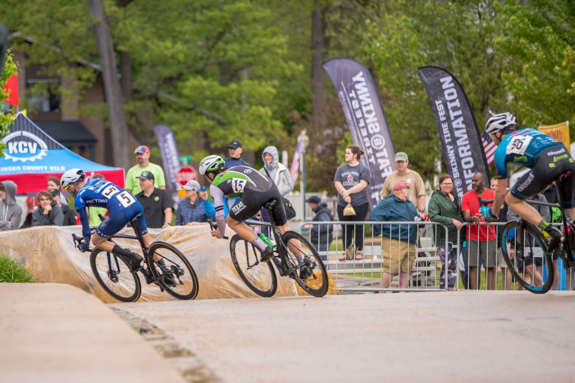 Cyclists compete in a race, leaning into a turn, with spectators watching behind barriers, flags, and banners in the background amidst trees.
