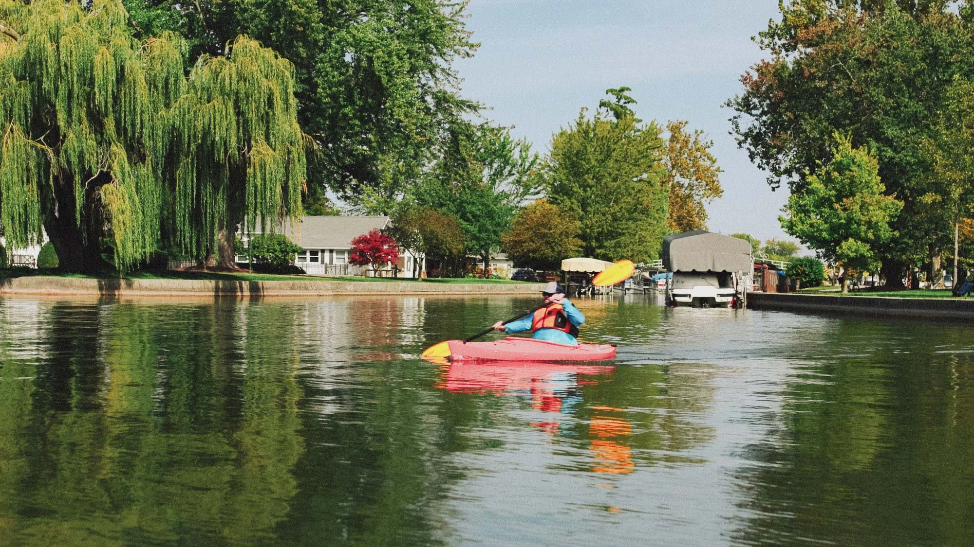 A person kayaking on a calm river, surrounded by lush trees with green and some orange leaves, and houses along the riverbank in the background.