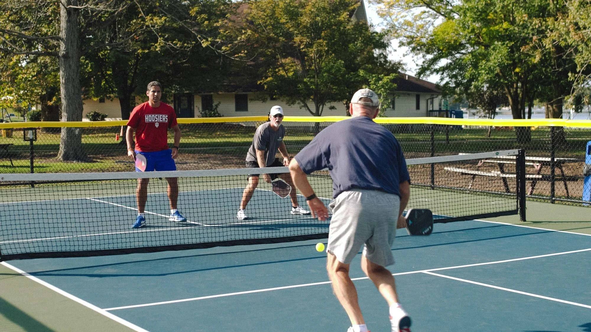Three men playing tennis on an outdoor court, one is about to hit the ball, with trees and residential houses in the background.