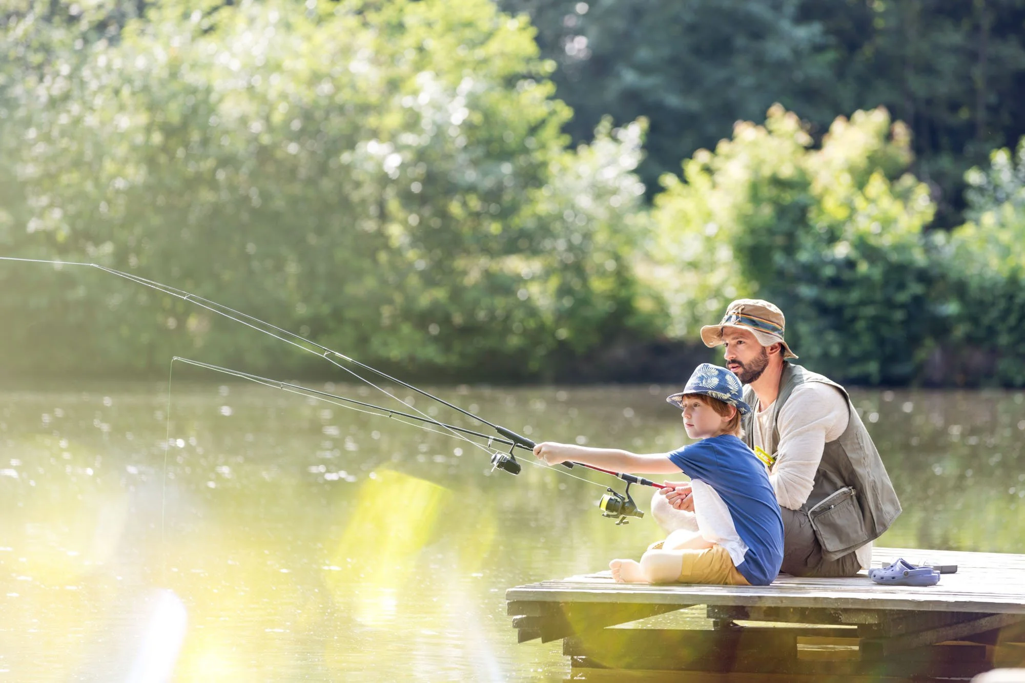 A man and boy fishing together on a dock by a lake, with trees in the background during daytime.
