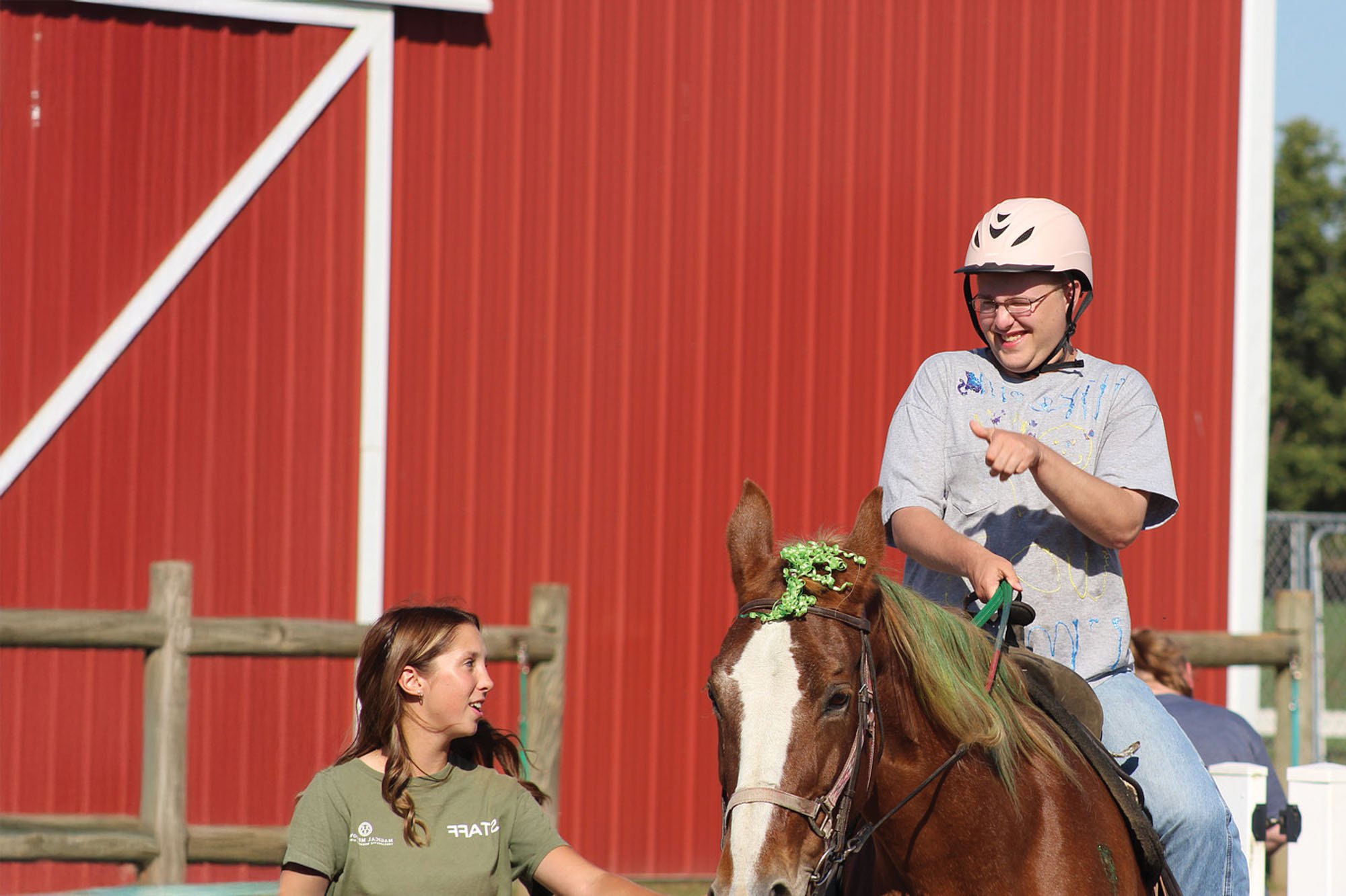 A young man wearing a white helmet and glasses, riding a horse with a decorated green mane, is smiling and pointing. A young woman with long brown hair, wearing a green T-shirt, is looking up at him. In the background, there is a red barn with white 