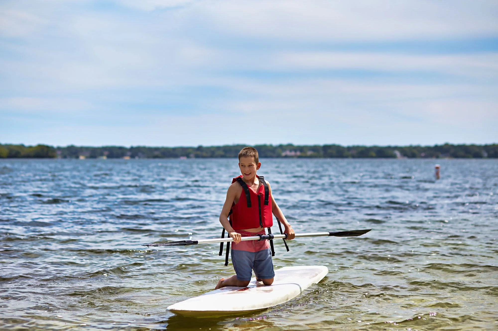 A boy standing on a paddleboard in water, wearing a red life jacket and holding a paddle, with a scenic lake and blue sky in the background.