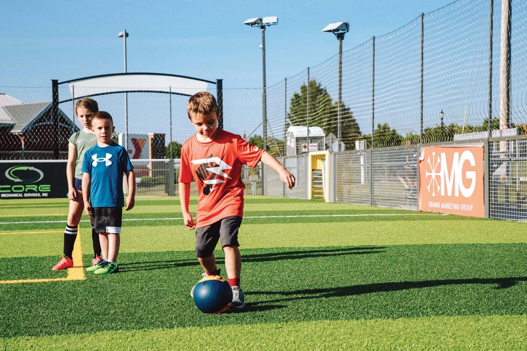 Three kids playing soccer on a field, with one boy in an orange shirt kicking a ball and two children in shirts waiting behind him.