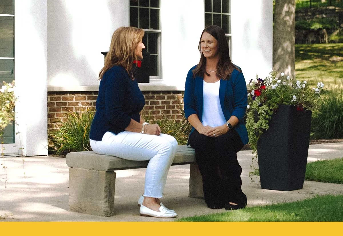 Two women having a conversation outdoors, seated on a stone bench near a building with brick and siding, surrounded by greenery and flowers.