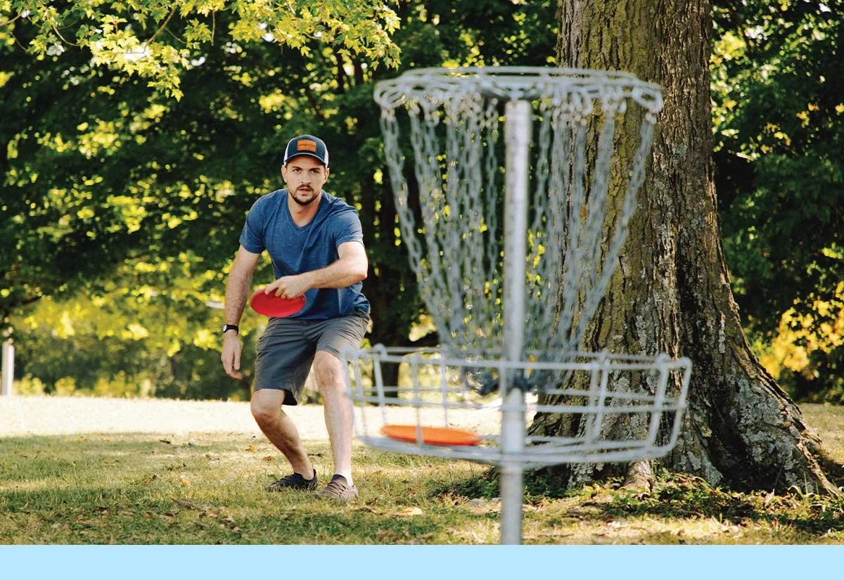 A man playing disc golf outdoors near a large tree, holding a red disc, with a metal disc golf basket in the foreground.