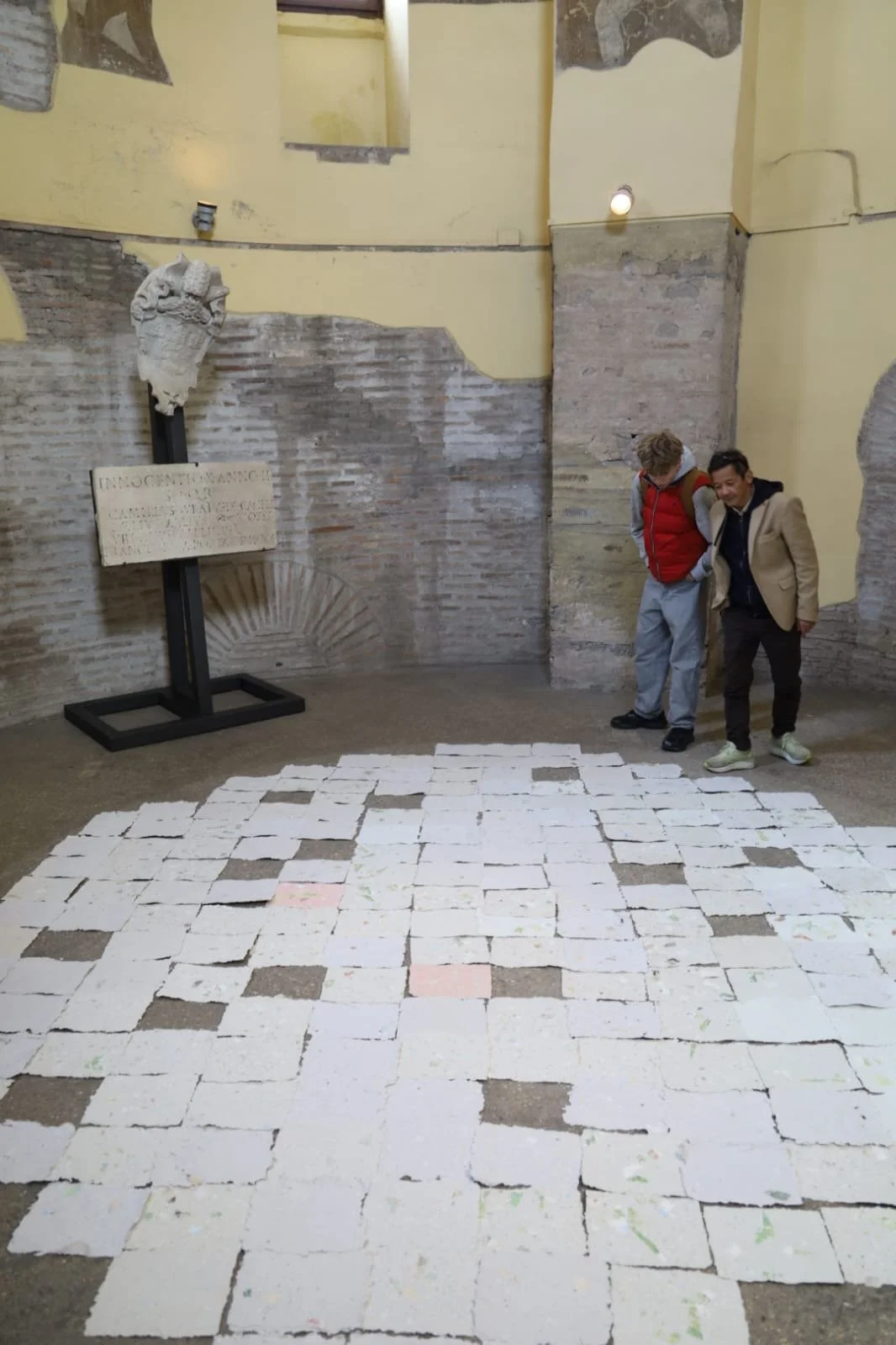 Two men standing inside a historical ruin or site, looking down at a floor covered with white paper squares arranged in a grid pattern. The background features brick and plaster walls, a sculpture bust on a stand, and a sign with Latin text.